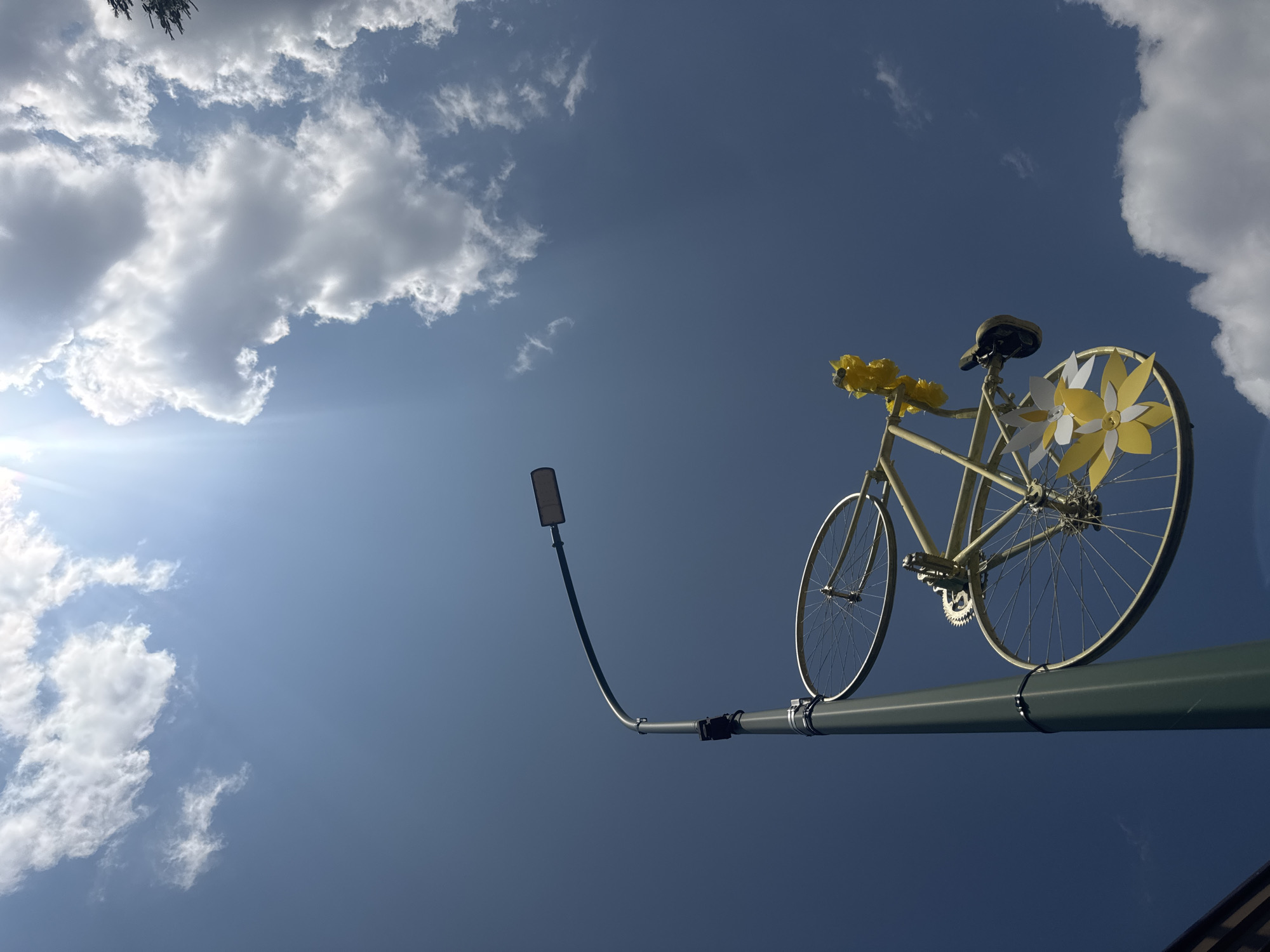 A bicycle, painted yellow and adorned with decorative yellow and white flowers, is mounted horizontally on a tall, dark green lamppost. The bicycle is positioned on the side of the lamppost, with one wheel above the other. A streetlight fixture is attached to the curved arm of the lamppost. The scene is set against a bright blue sky with scattered white clouds and the sun shining intensely from the upper left quadrant, creating a lens flare. The image appears to be taken from a low angle, looking upwards. While the prompt mentions Morbier, France, there are no specific visual cues in the image to confirm this location.