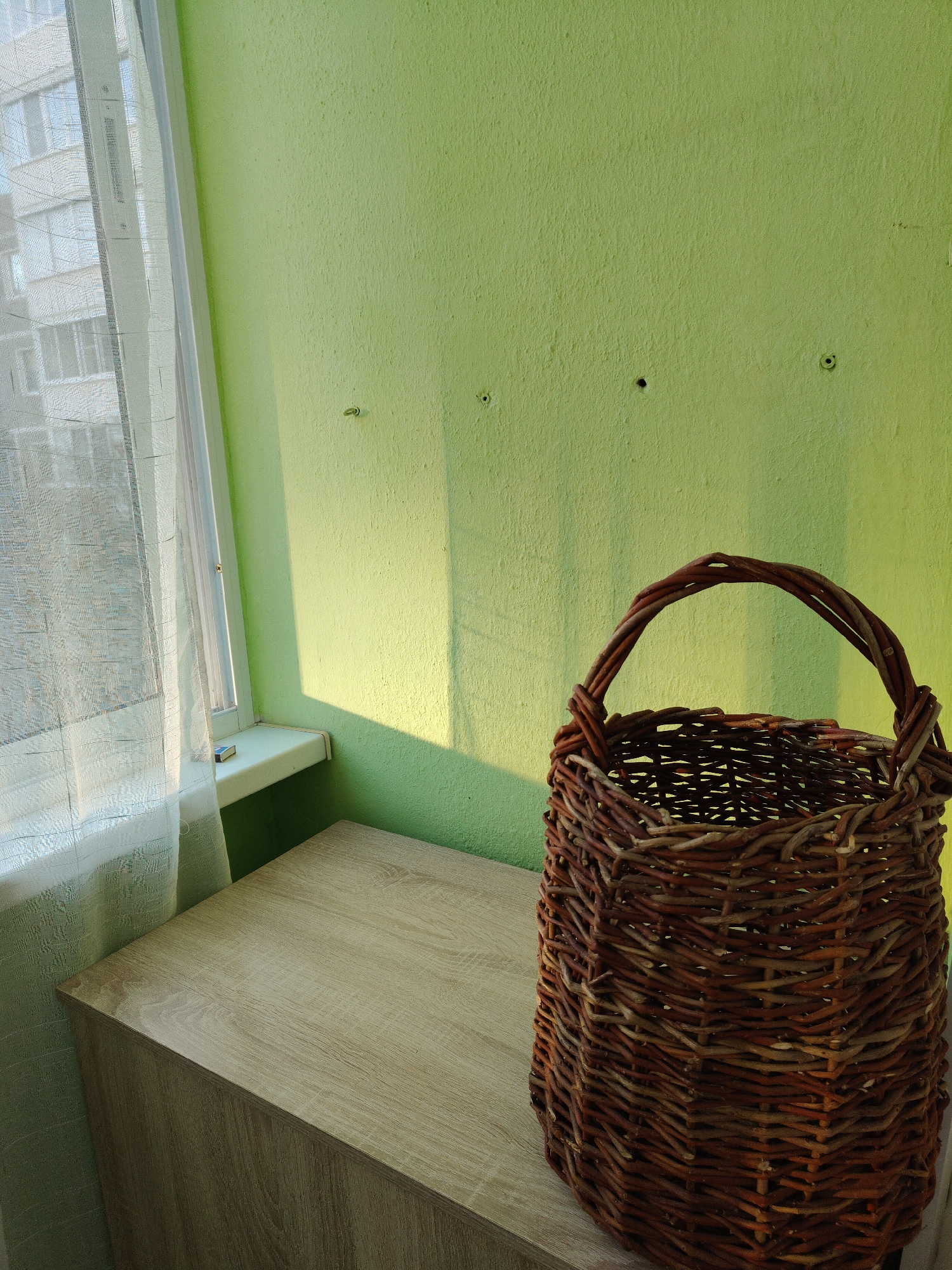 The scene is an interior shot of a room in Unknown, Unknown, featuring a woven wicker basket placed on top of a light wood-grain cabinet. The cabinet is positioned against a light green wall that has three drilled holes. Beside the cabinet is a window covered by a sheer white curtain that partially obscures the view of a building on the other side. A box of matches is visible on the window sill. The lighting suggests daytime conditions.