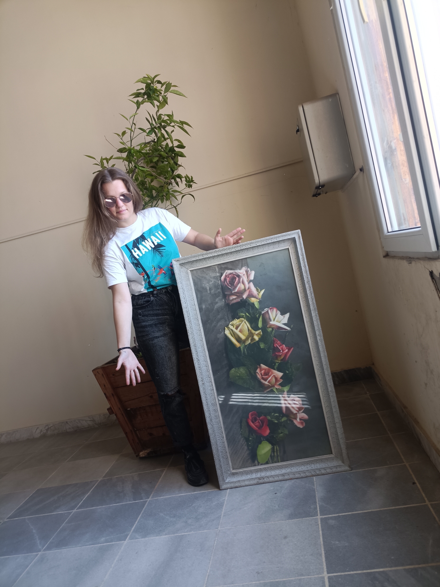 A young woman with long, light brown hair and rounded sunglasses poses indoors in Plovdiv, Bulgaria, alongside a large framed picture. She wears a white t-shirt with a blue "HAWAII" graphic depicting palm trees and surfers, dark distressed jeans, and black sneakers. Her body is slightly angled, with her left arm extended and palm open as if presenting, while her right arm is bent, resting near the top of the framed artwork. She is leaning against a rustic wooden crate or box.
The prominent object is the vertically oriented framed picture, featuring a vibrant bouquet of pink, yellow, and red roses in a vase against a dark, textured background. The frame is a distressed light grey. Behind the woman, a leafy green plant adds a touch of nature. The setting features plain light beige walls and a grey tiled floor with visible grout lines. Natural daylight enters from a white-framed window on the right, near which a small wall-mounted utility box is visible. The overall scene suggests a casual, well-lit indoor or semi-outdoor space, possibly a balcony or hallway. The time of day is daytime, indicated by the bright natural light. The woman's expression is neutral and calm.