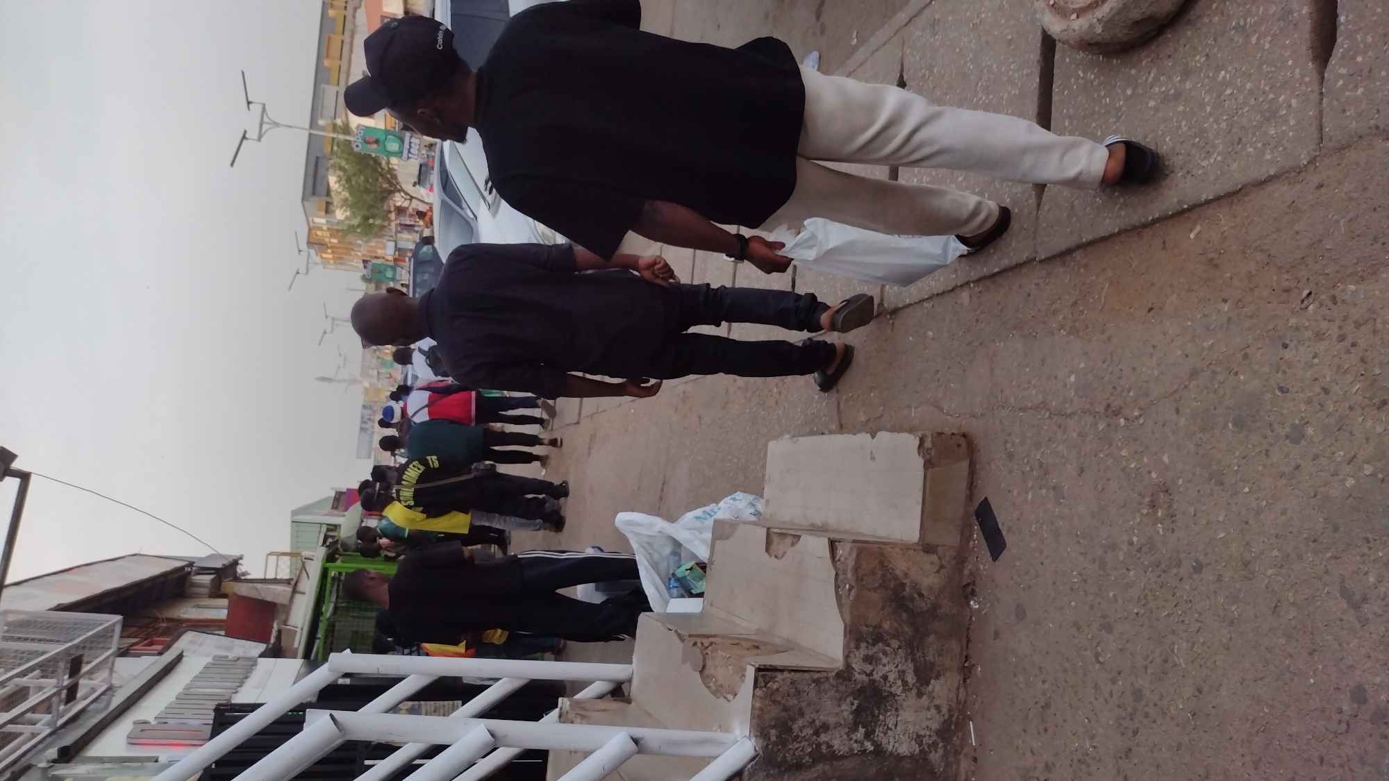 A sidewalk scene in Jos, Nigeria, features several individuals walking along a paved area. In the foreground, two men wearing black shirts and dark trousers (one with light trousers) are observed from the side, with one carrying a white plastic bag. A group of people, some wearing colorful clothing, are further down the sidewalk. The built environment includes buildings and street infrastructure such as lampposts. Debris and construction materials are scattered near the sidewalk's edge.
