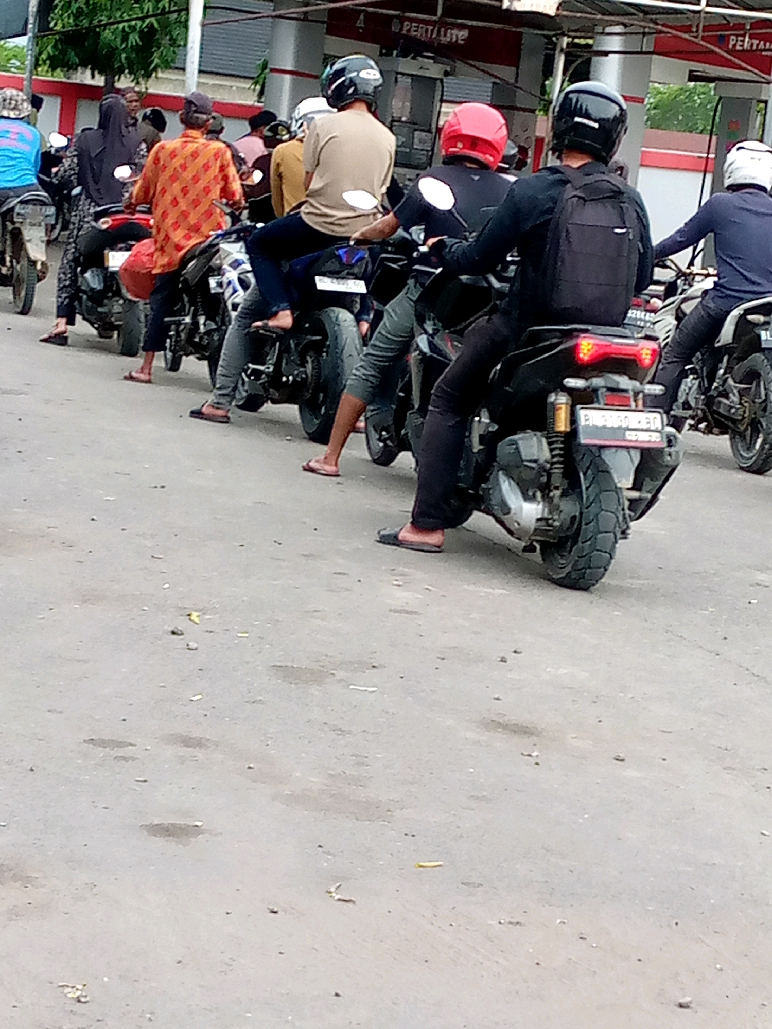 The image depicts a line of motorcyclists at what appears to be a gas station, likely in Keudebayu, Indonesia, given the context. The scene is captured from a slightly elevated, rear-facing perspective, focusing on the backs of several individuals and their motorcycles.
In the foreground, a motorcyclist wearing black pants and a black backpack is seated on a dark-colored motorcycle. They are wearing a black helmet. To their right, another motorcyclist in a dark shirt and black pants is visible from the back, also wearing a black helmet. Further to the right, a third motorcyclist wearing a dark blue shirt and dark pants is also seen from the back, wearing a white helmet.
Behind the foremost motorcyclist, there are several other people on motorcycles. One individual is wearing an orange and red patterned shirt and a dark cap, seated on a motorcycle. To their left, a person in a black headscarf is on a motorcycle. Another motorcyclist in a blue shirt is further to the left. The background includes a building structure with red and white accents, possibly part of the gas station infrastructure, and some trees. The ground is paved and appears somewhat worn. The overall impression is a mundane scene of people waiting in line on their motorcycles.