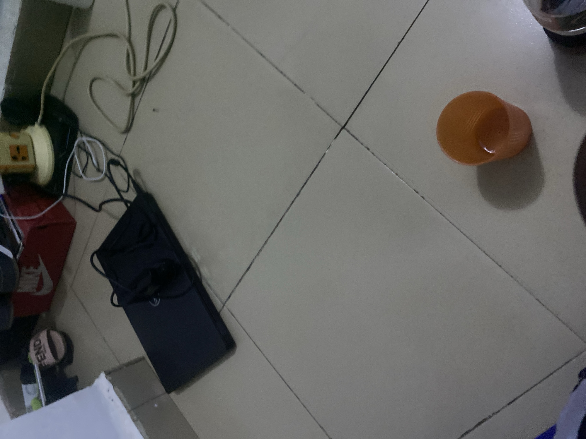 This is a close-up, eye-level shot of a tiled floor in Howayi, Nigeria. The scene depicts a collection of everyday objects. In the foreground, a small, orange plastic cup with ribbed sides is visible. To the left, a black laptop computer is placed on the floor, with its lid closed. A black mouse and its cord are resting on top of the laptop. Further back, several power cords, one white and one beige, are tangled on the floor. Behind the laptop, a red Nike shoebox is partially visible. Next to the shoebox, a light-colored slide sandal with the word "FENDI" in black letters is on the floor. The background consists of a white wall and a yellow electric fan. The overall impression is of a casual, perhaps lived-in, domestic environment.