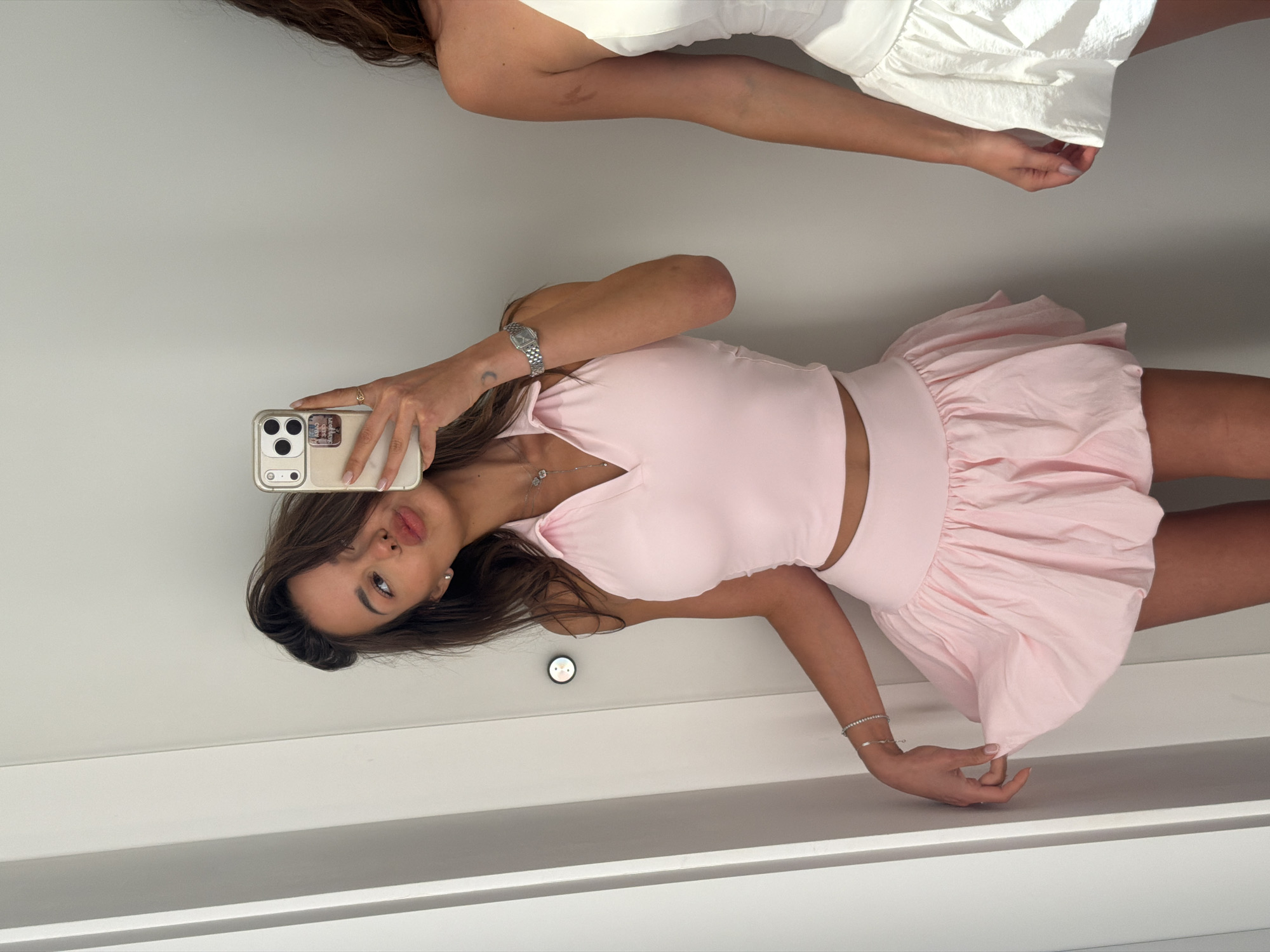 A young woman with dark hair and light skin is taking a selfie in a fitting room. She is wearing a pale pink halterneck crop top and a matching bubble skirt. She is pouting her lips and holding her phone up to take the picture. There is a white outfit on a mannequin to her right. The fitting room has white walls and a white door. The woman has a watch on her left wrist and a bracelet. The environment is well-lit, likely an indoor space such as a clothing store or a home dressing room. It is not possible to determine the location in the city of Saint Petersburg, Russia, based on the provided image.