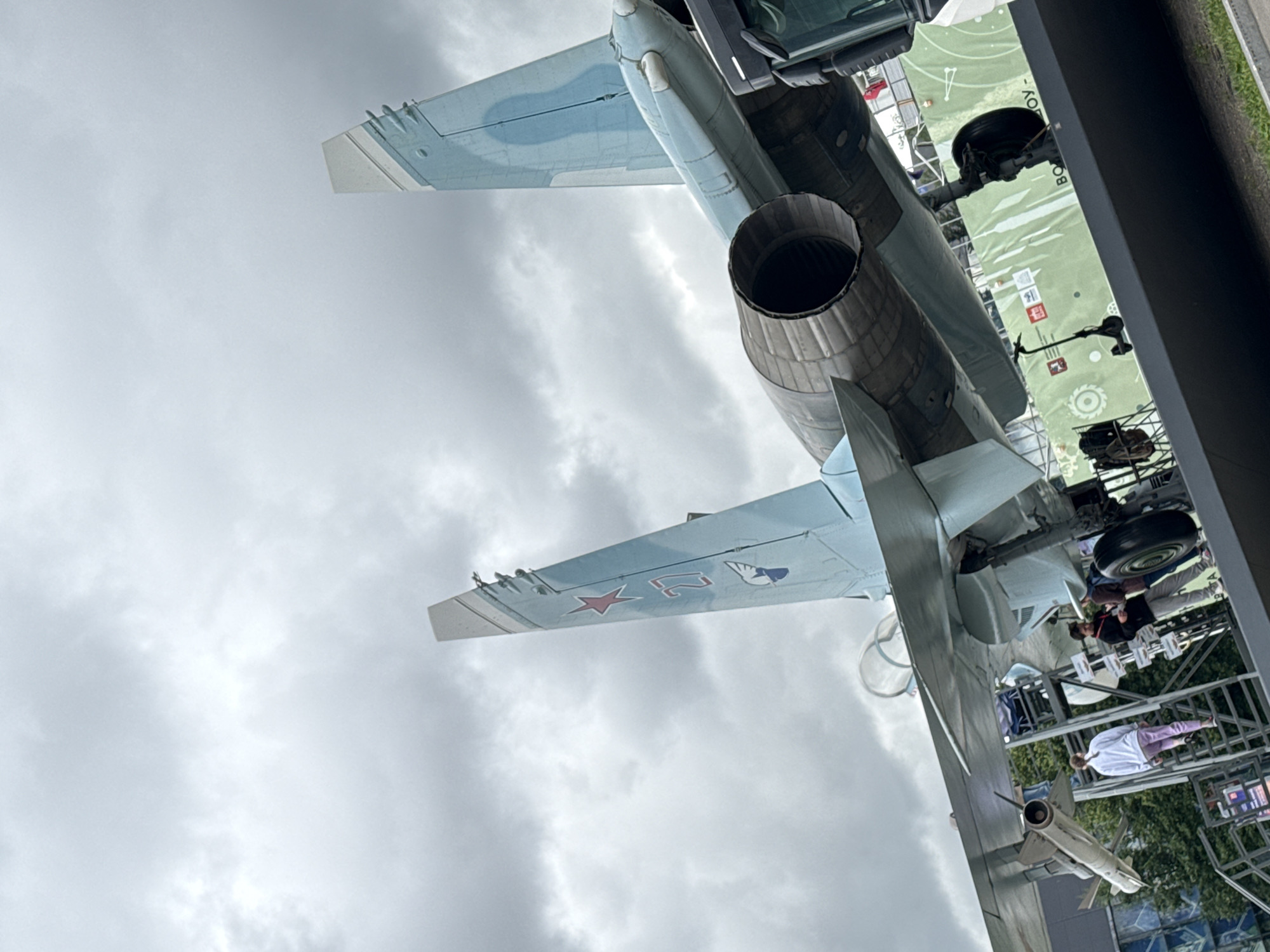 This is a low-angle, outdoor, waist-level shot of the underside and rear of a fighter jet, likely on display at an aviation museum or airshow in Moscow, Russia. The jet is predominantly light blue with some darker camouflage patterns on its wings. It features a prominent engine exhaust at the rear. The sky is overcast and cloudy, suggesting a cool or temperate day.
Several people are visible in the background, appearing to be visitors. Some are walking around the jet, while others are looking up at it. One person is wearing a light-colored jacket and purple pants. Another person in the background is wearing a dark jacket and appears to be speaking with someone. The jet is supported by a metal structure, and a green banner with white text and graphics is visible behind it. The text on the banner is partially obscured but includes Cyrillic characters. A missile is attached to the underside of the jet. The overall impression is of a public viewing of a military aircraft.