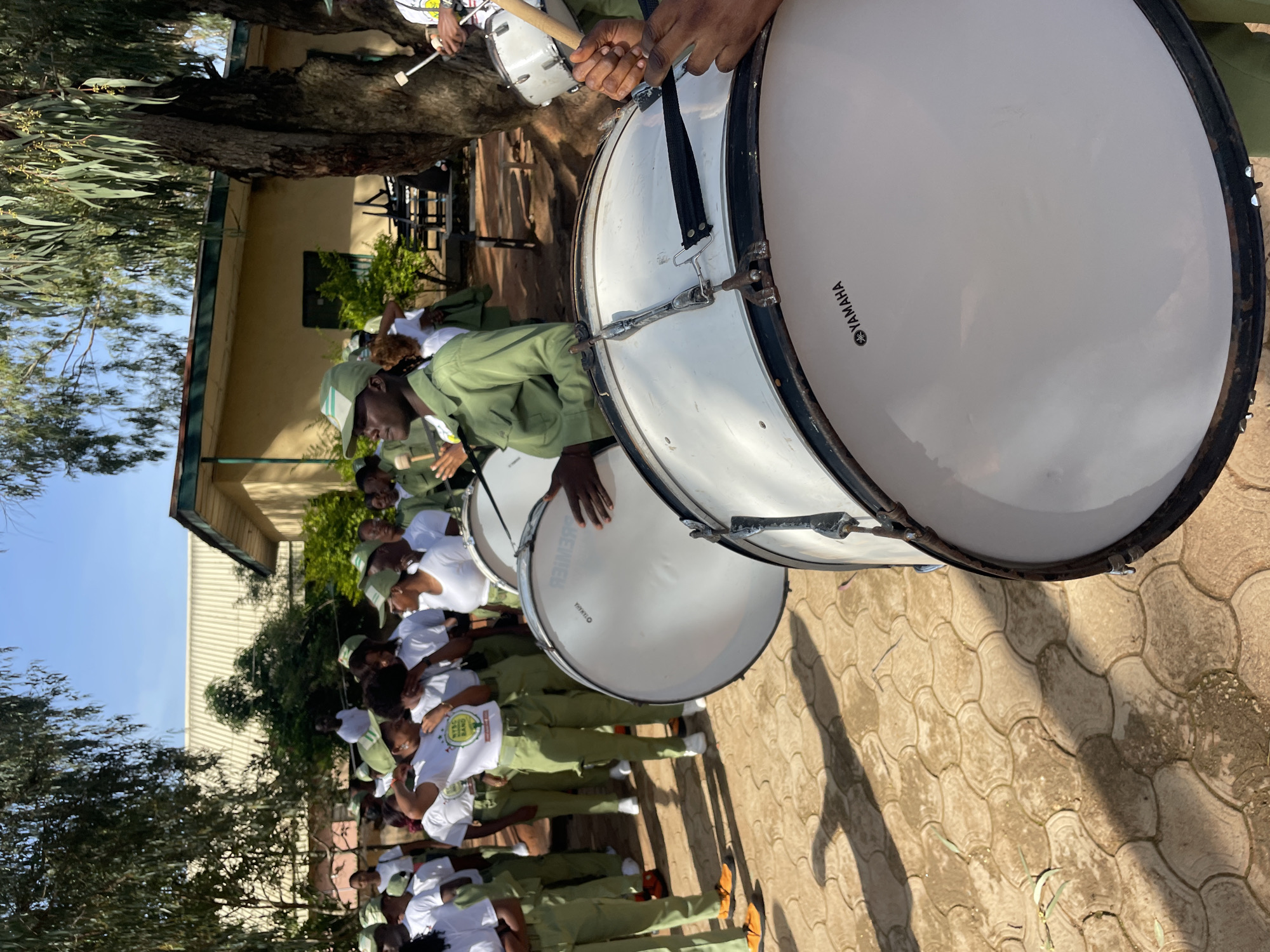 In Jos, Nigeria, a group of individuals, likely uniformed youth corps members, are gathered outdoors, preparing to play drums. They are dressed in matching green uniforms and caps, with some wearing white t-shirts with inscriptions, one of which reads "NYSC BAND." The setting appears to be a school or administrative compound, with a building visible in the background. Two large bass drums, branded "YAMAHA," are prominently featured in the foreground, with drumsticks held by unseen individuals, suggesting they are about to be played. Other members of the group are standing around, some interacting, and others looking towards the drums. The ground is paved with interlocking stones, and trees provide shade. The scene is illuminated by natural daylight, casting shadows on the ground.