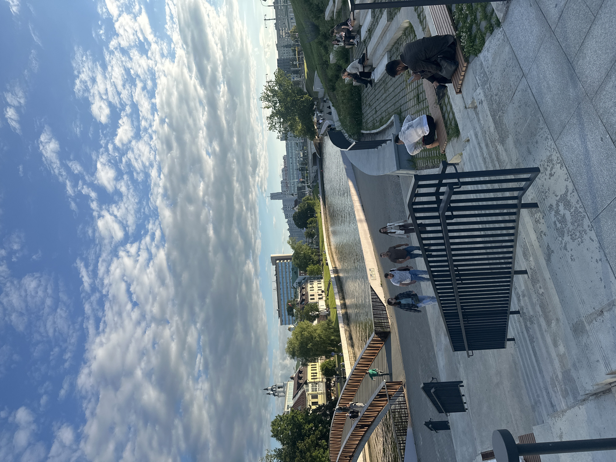 This image shows a wide view of a modern city park and riverfront in Minsk, Belarus. In the foreground, there are wide concrete stairs with black metal railings, leading down to a paved pathway. Several people are descending the stairs, and one person is sitting on a bench on the right side of the frame, looking at their phone. Beyond the stairs, a curved river embankment is visible, with a pathway and grassy areas. People are seen walking and sitting on benches along the embankment. A pedestrian bridge arches over the river in the mid-ground, with several individuals walking across it. The river itself is calm, reflecting the sky. On the far side of the river, a line of buildings with diverse architecture, ranging from older, traditional structures to modern high-rises, forms the city skyline. Trees are scattered throughout the park and along the riverbank. The sky is a prominent feature, filled with a mix of blue sky and dramatic, fluffy white and grey clouds, suggesting a dynamic weather day. The overall atmosphere is one of leisure and urban revitalization.