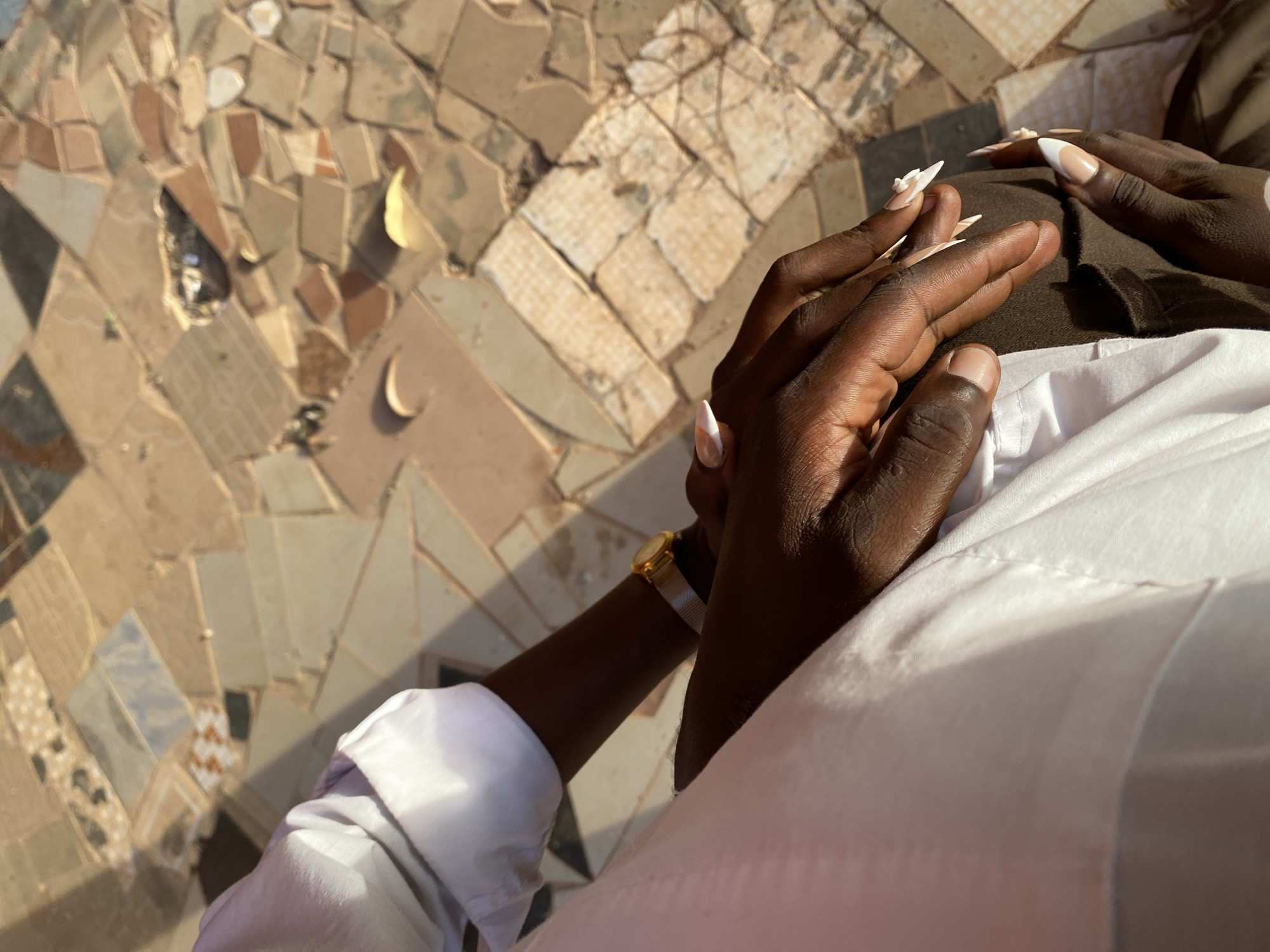 A close-up, high-angle shot shows a person seated on a mosaic surface. The mosaic features light tan, beige, and brown tiles. The person has dark skin and is wearing a white collared shirt with the sleeve rolled up. A gold wristwatch is visible on the left wrist. The person's hands are clasped together, with manicured nails, one hand on top of the other. The left hand has a white floral design. The person's hands rest on brown fabric. This image was not taken in the city of Vom, Nigeria.