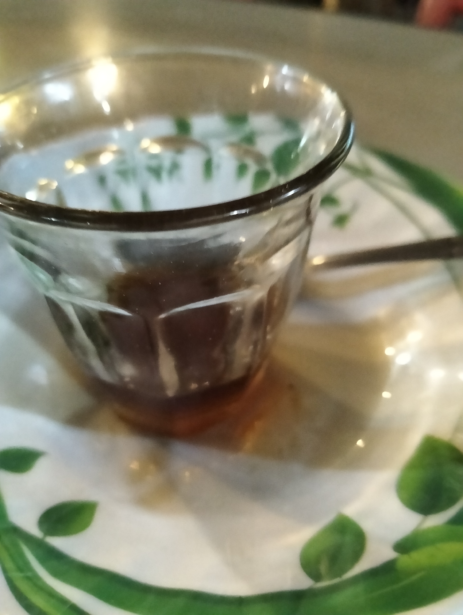 A close-up, shallow depth-of-field shot shows a clear glass cup filled with a dark liquid, possibly coffee or tea, resting on a white saucer with a green leaf pattern. The saucer also appears to have a subtle floral design. A silver spoon is partially visible, placed to the right of the cup, resting on the saucer. The background is blurred, with hints of warm lighting and a reflective surface suggesting an indoor setting. The image does not provide any identifiable features or context to suggest its location in Payagaboh, Indonesia.