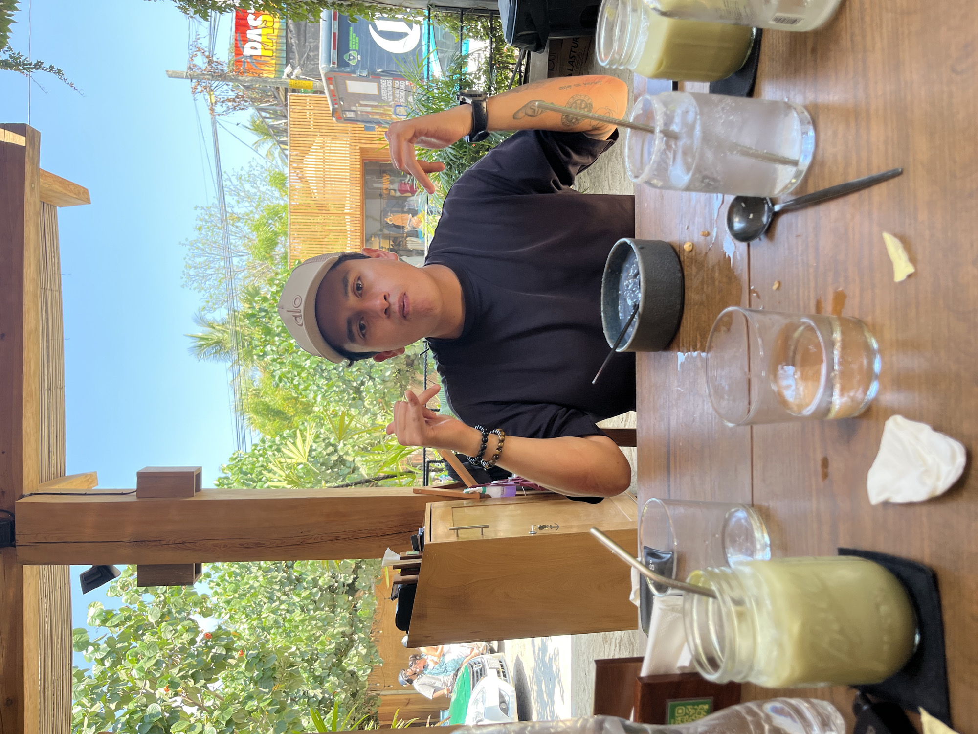 The image captures a man sitting at a wooden table, likely at an outdoor cafe, with several empty glass containers and jars, some with straws or spoons still inside. He is wearing a dark T-shirt, a tan cap with "ALO" on the front, and bracelets. He is making a playful gesture with his hands.
The background shows a street scene with trees, buildings, and a vehicle with signage. Based on visual cues, the location is identified as Puerto Escondido, Mexico.