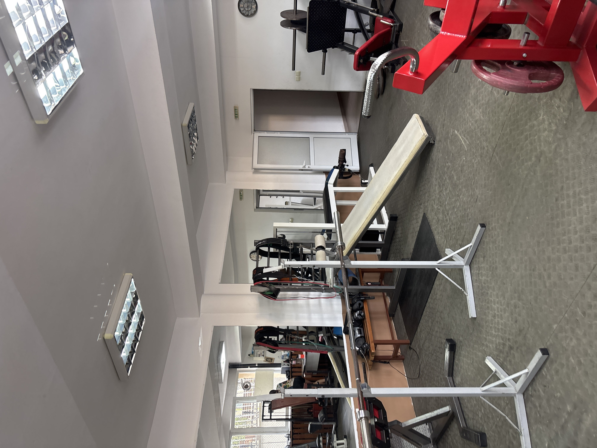 This image depicts the interior of a gym, likely located in Zlatograd, Bulgaria, judging by the available information. The gym features various exercise machines, including a bench press with adjustable supports, a leg press machine in the upper right, and other weightlifting equipment. Mirrors line one wall, reflecting the gym equipment and the interior. The floor is covered with a rubberized, textured surface. The lighting is provided by fluorescent ceiling lights. A doorway with a glass panel is visible in the background. The gym appears to be empty of people.