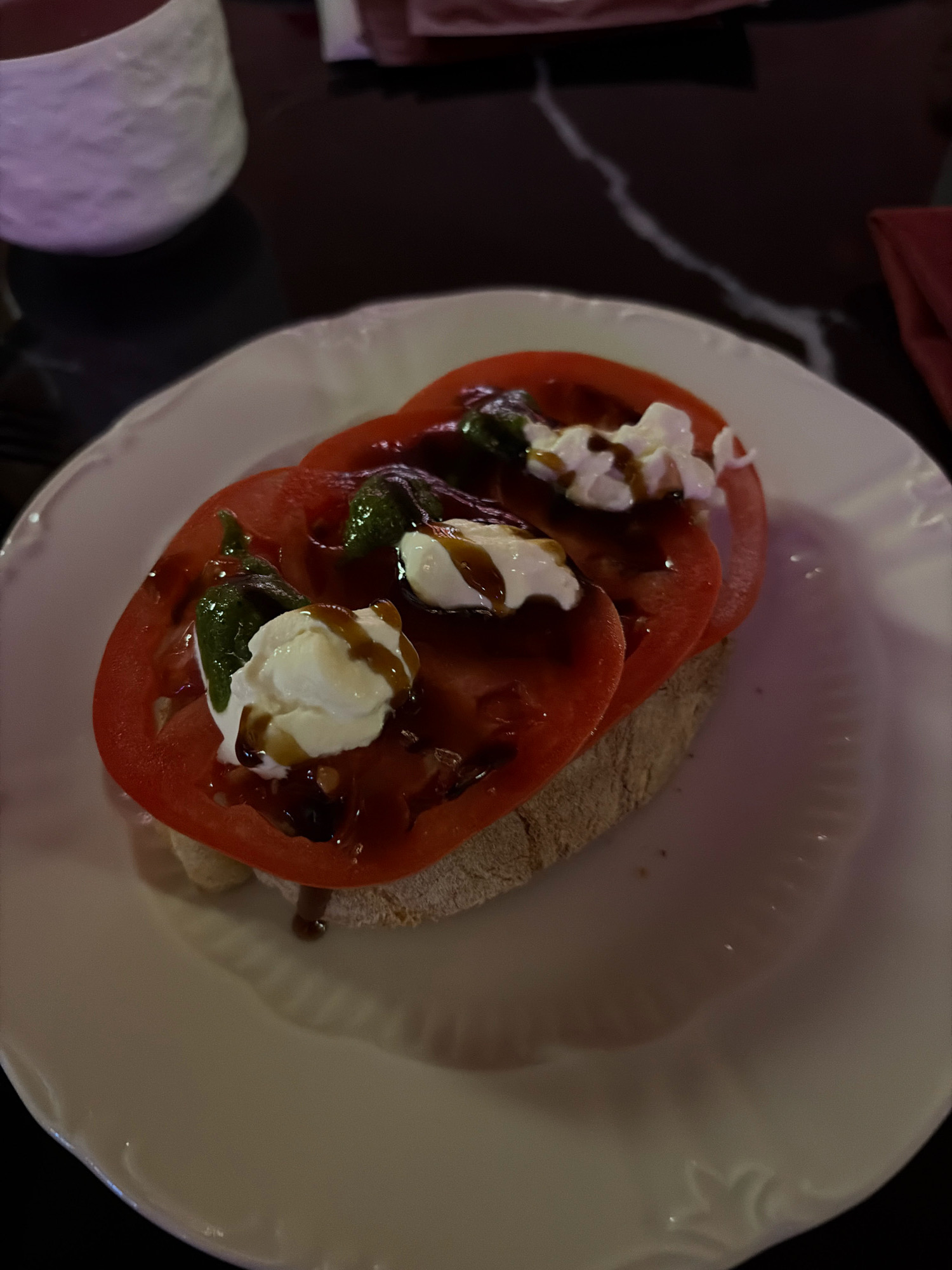 A close-up, eye-level shot shows a piece of bruschetta on a white plate. The bruschetta is topped with two thick slices of ripe tomato, fresh basil leaves, dollops of white cheese, and a drizzle of dark balsamic glaze. The bread appears to be a rustic, whole-grain variety. The white plate has a slightly scalloped edge and is set against a dark, out-of-focus background. To the left of the plate, a portion of a white textured ceramic cup is visible. The lighting is dim, creating shadows and highlighting the glossy texture of the tomatoes and glaze. There are no people present in the image. The scene is indoors, likely in a restaurant setting. The location is not specified in Astana, Kazakhstan.