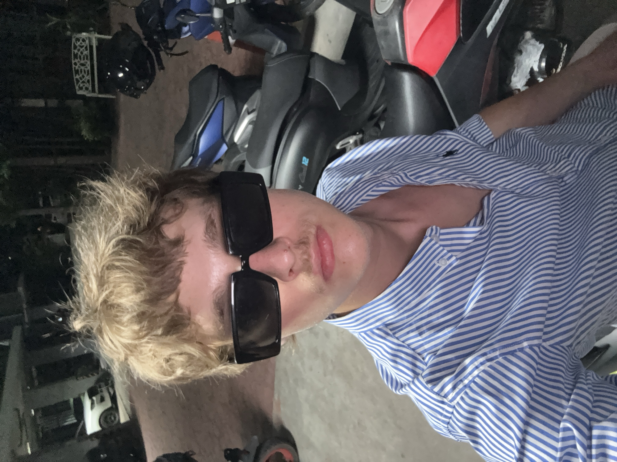 The media file shows a close-up, high-angle, portrait-style shot of a young person with blonde, tousled hair and a thin mustache, wearing large, black, rectangular sunglasses. They are also wearing a blue and white striped button-down shirt. The person is looking directly at the camera with a neutral expression.
Behind the subject, several motorcycles are visible, suggesting an outdoor setting, possibly a parking area or a street. The lighting is dim, indicating that it is likely evening or nighttime. The background is out of focus but shows more motorcycles and some foliage, hinting at a warm climate.
The overall mood is casual and possibly a bit candid, as the subject is wearing sunglasses indoors or at night. No specific activity is clearly depicted, but the presence of motorcycles implies that transportation is relevant to the scene. There are no visible text elements in the image. The location context is Rawai, Thailand.