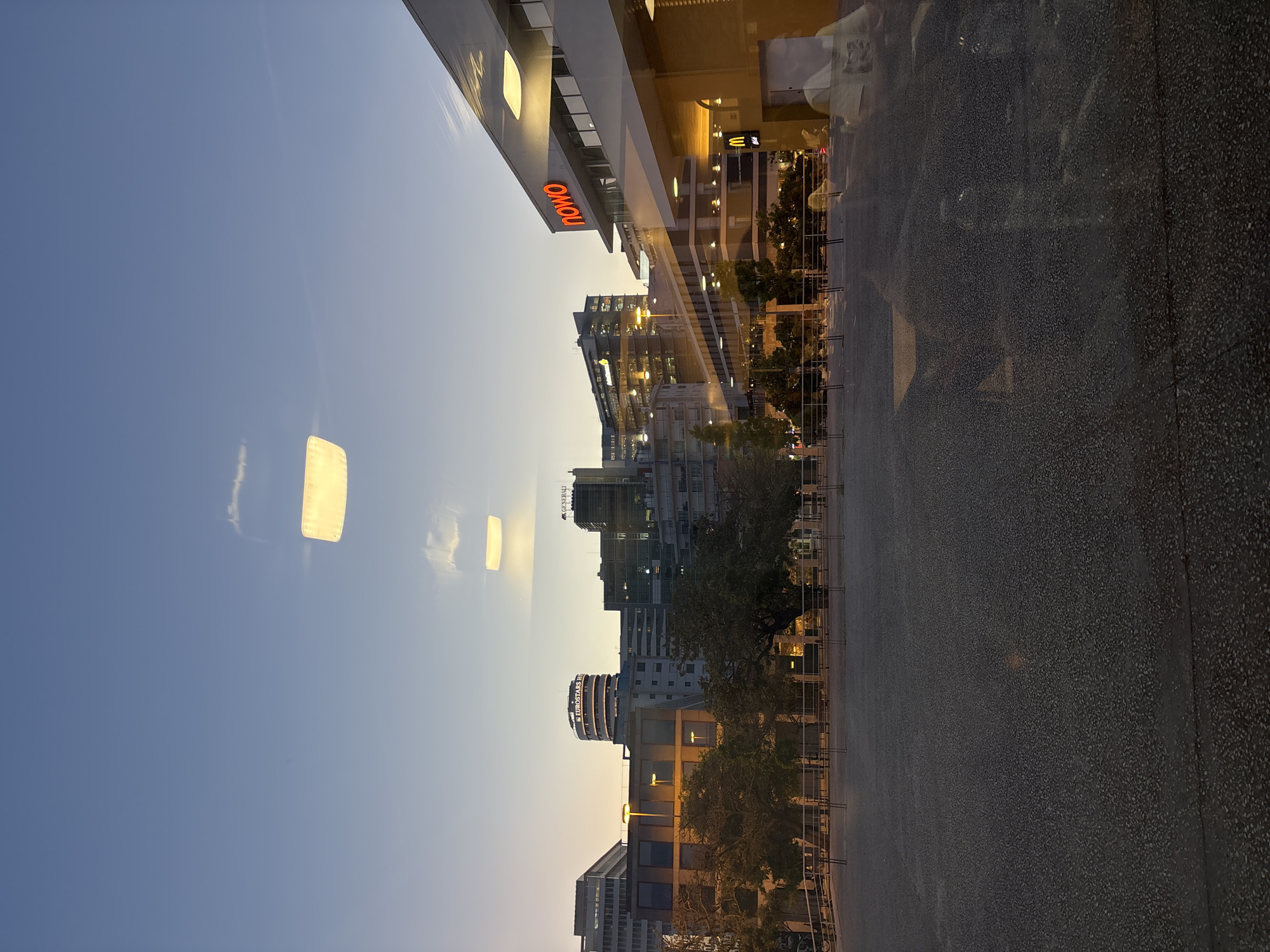 The image depicts a view in Lisbon, Portugal, captured through a glass surface, likely a window. The scene is oriented vertically. The sky is visible at the top left, transitioning from blue to a lighter hue as the sun sets or rises. The reflections on the glass show city buildings, with some recognizable details like a building with a red "NOWO" sign and a "McDonald's" sign further down the street. The "Eurostars Hotel" is also visible. The lower right part of the image captures the sidewalk or ground in front of the viewer. Trees line the street, with metal fencing visible. The lighting suggests either dusk or dawn, with warm tones cast on the buildings. There are no people visible in the primary view, although reflections might suggest their presence in the surrounding environment.