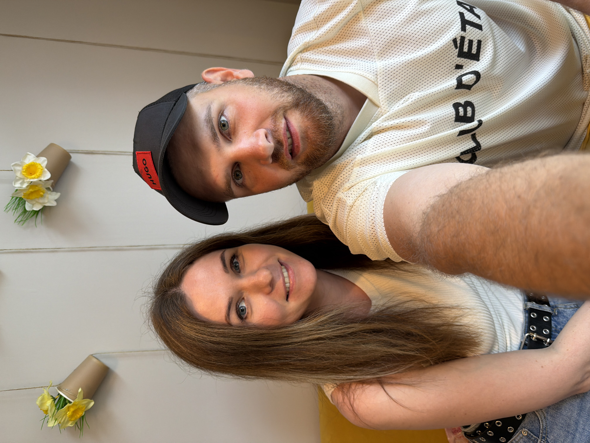A man and a woman are taking a selfie. The man is wearing a black baseball cap with a red "HUGO" logo and a light-colored mesh shirt with black lettering that appears to read "CLUB D'ETA". He has a beard and light-colored eyes. The woman has long, light brown hair and blue eyes. She is wearing a light-colored ribbed top and blue jeans with a black belt. They are sitting side-by-side, with the man holding the camera. In the background, there is a light-colored wall decorated with two arrangements of artificial yellow and white flowers in brown paper cups, hanging by twine. A yellow sofa is visible to the left of the woman. The scene appears to be indoors, possibly in a cafe or home. There are no recognizable landmarks or features that would indicate the specific location within Chaikovsky, Russia.