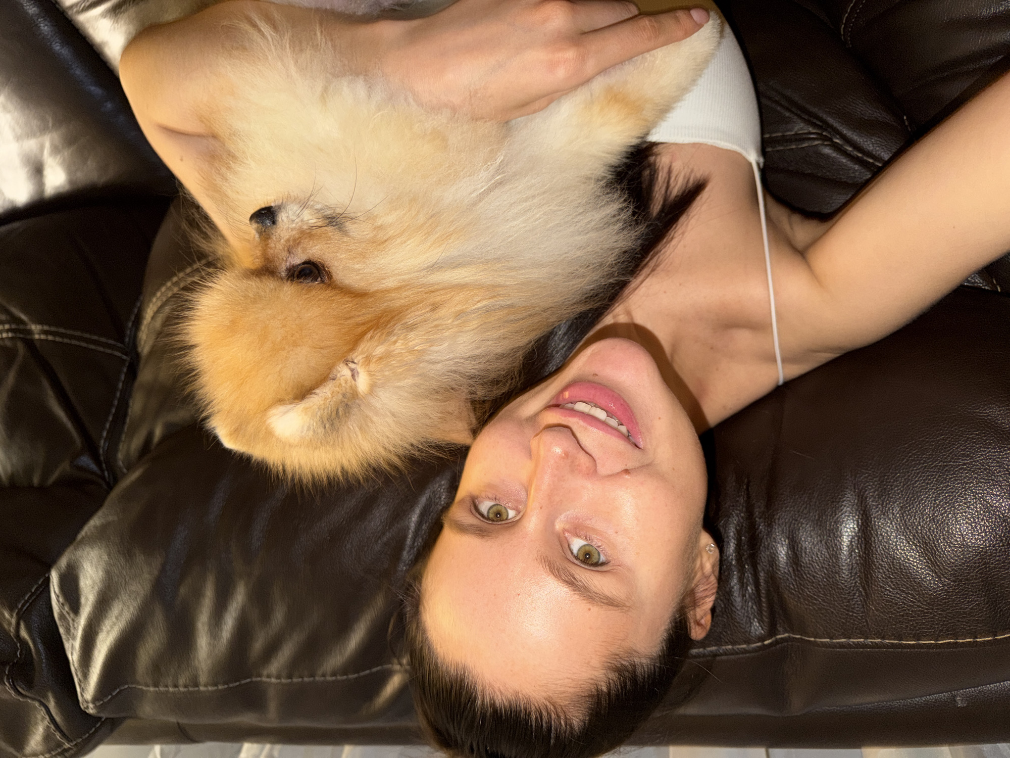 An overhead shot captures a woman and a small dog relaxing together on a dark brown leather couch in what appears to be an indoor setting in Khimki, Russia. The image is rotated, showing the woman's head positioned towards the bottom right of the frame, smiling slightly and looking directly at the camera. She has green/hazel eyes and dark hair tied back, and is wearing a simple white spaghetti-strap top.
A fluffy, golden-orange and cream-colored dog, likely a Pomeranian, is nestled on her chest and shoulder area, resting on its back with its head near the woman's neck. Its black nose and dark eyes are visible. The woman's left arm extends upwards, gently resting near the dog. The couch, visible with its characteristic stitching and creased texture, fills the majority of the background, suggesting a comfortable and relaxed environment. The overall mood is calm and affectionate, depicting a moment of companionship between the woman and her pet. No visible text is present in the image.