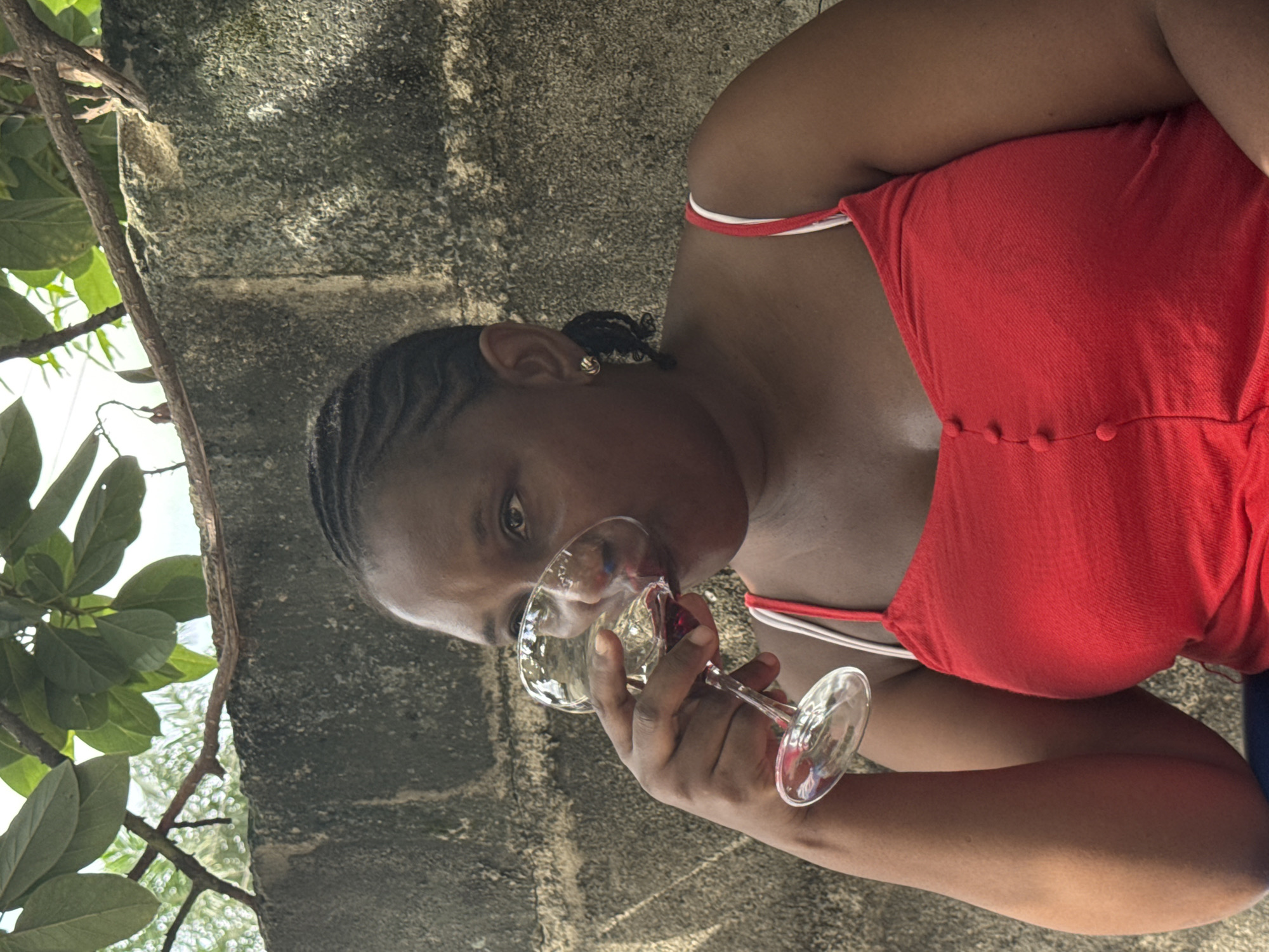 A woman with braided hair, wearing a red top with white straps, is drinking from a wine glass. She is positioned in front of a weathered, grey wall, possibly made of concrete blocks. To the left of the wall, there's greenery with leaves and branches. The location is inferred to be in Ado, Nigeria.