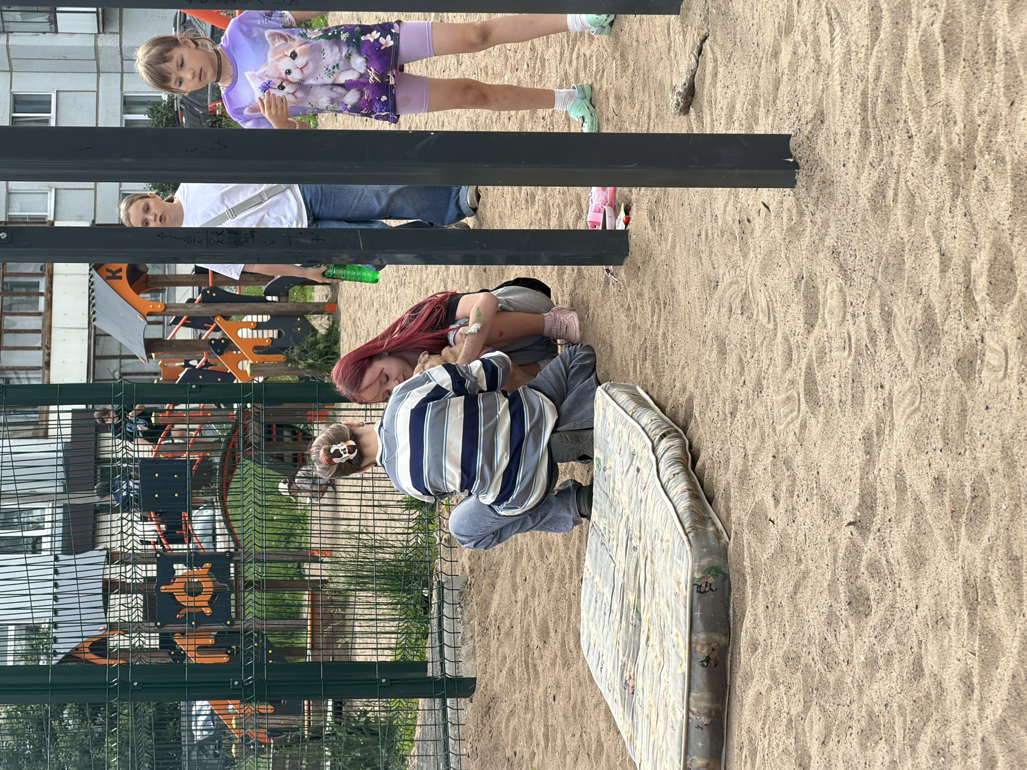 In a sandy playground in Chaikovsky, Russia, three girls are present. A girl with red hair is squatting, holding a small dog. Another girl, wearing a striped shirt and jeans, is also squatting and looking at the dog. A third girl, wearing a white t-shirt and jeans, stands to the left. To the right, another girl in a purple t-shirt with cats is visible. A discarded mattress lies in the sand in the foreground. In the background, a colorful playground structure and apartment buildings are visible.