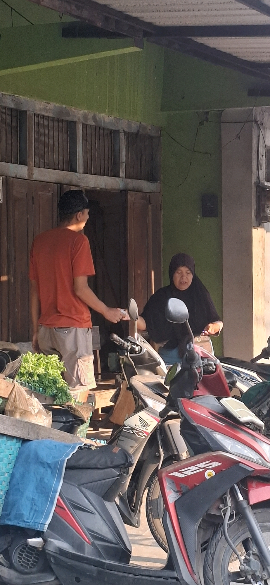In Desa Dibal, Indonesia, a man in a red t-shirt and grey cargo shorts stands in front of a wooden building with a green wall. He is handing a small white object, possibly money or a receipt, to a woman wearing a black hijab and black clothing. The woman is seated on a motorcycle. In the foreground, several motorcycles are parked. To the left, a crate filled with green leafy vegetables is visible, along with a blue cloth draped over a motorcycle seat. The scene appears to be an outdoor transaction occurring on a street or in front of a shop.