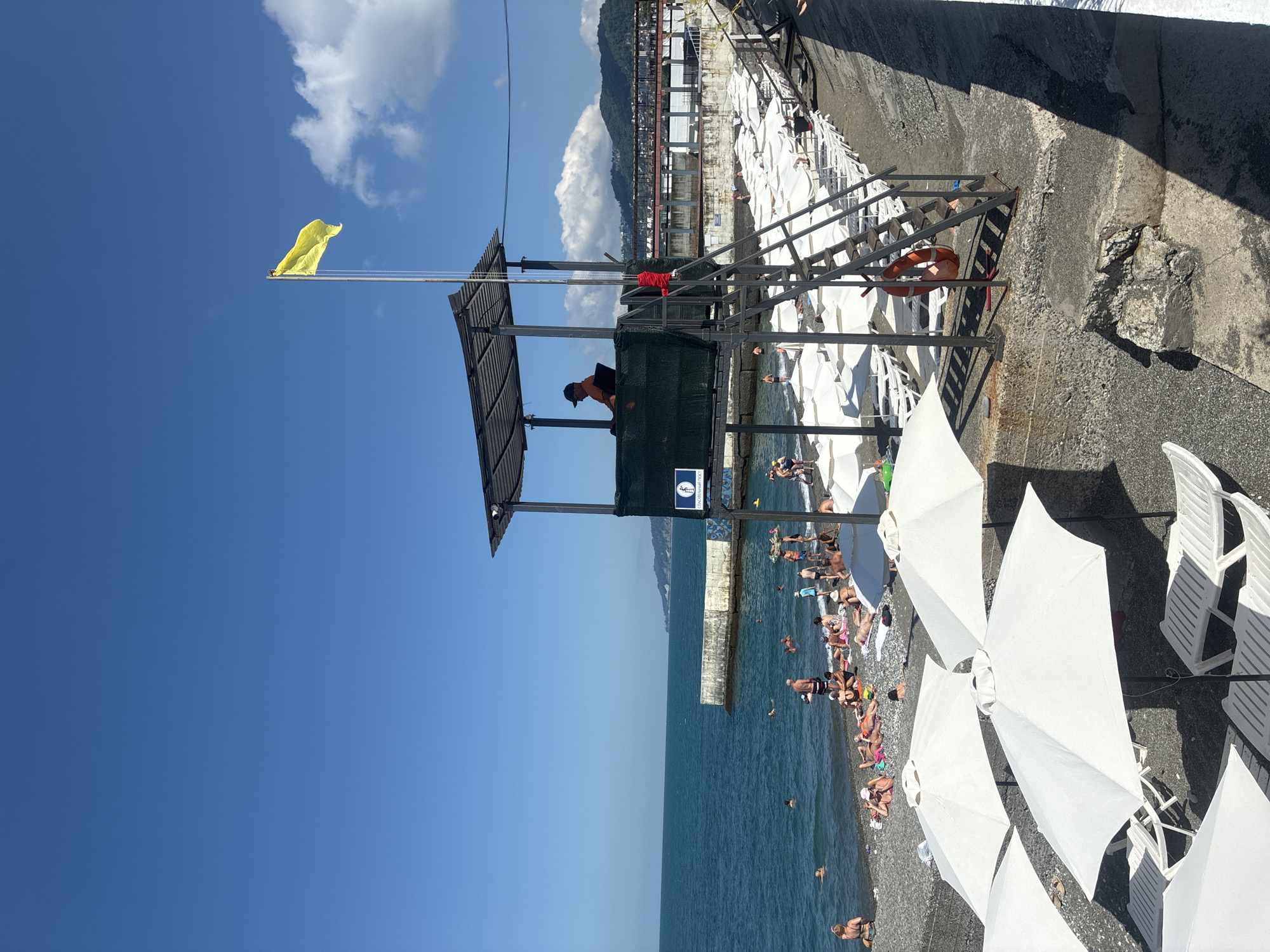 The image depicts a sunny day at a pebble beach in Sochi, Russia. A lifeguard tower with a yellow flag stands prominently in the foreground. Several beachgoers are visible, some swimming in the clear blue water, while others are sunbathing on lounge chairs under white umbrellas. A concrete breakwater extends into the sea, and in the background, buildings and lush green hills are visible. The overall scene suggests a typical summer day at a resort destination.