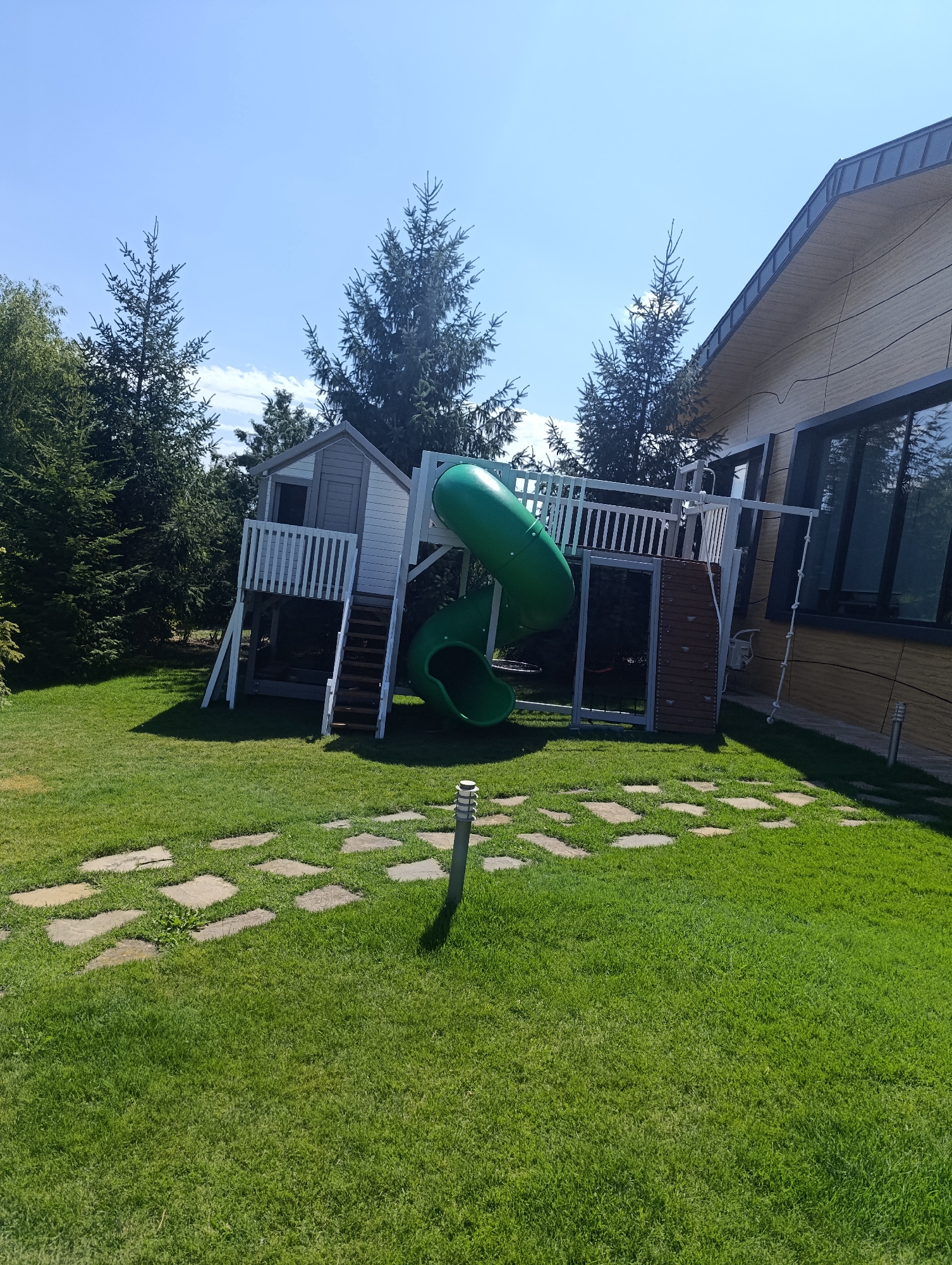 The image depicts a backyard scene on a clear, sunny day. In the foreground, a well-maintained lawn is visible, with a path of square stone pavers leading towards a grey garden lamp. Behind the lawn, a modern wooden house with a dark metal roof and large windows is situated. To the left of the house, a children's play structure is present. This structure includes a small white and grey playhouse elevated on stilts, accessed by wooden stairs. Adjacent to the playhouse is a bright green, curved slide. The play structure is surrounded by lush green trees, some of which are tall coniferous trees. The overall impression is of a residential property in a semi-rural or suburban setting, likely in Bulatselivka, Ukraine. There are no people or animals visible in the image.
