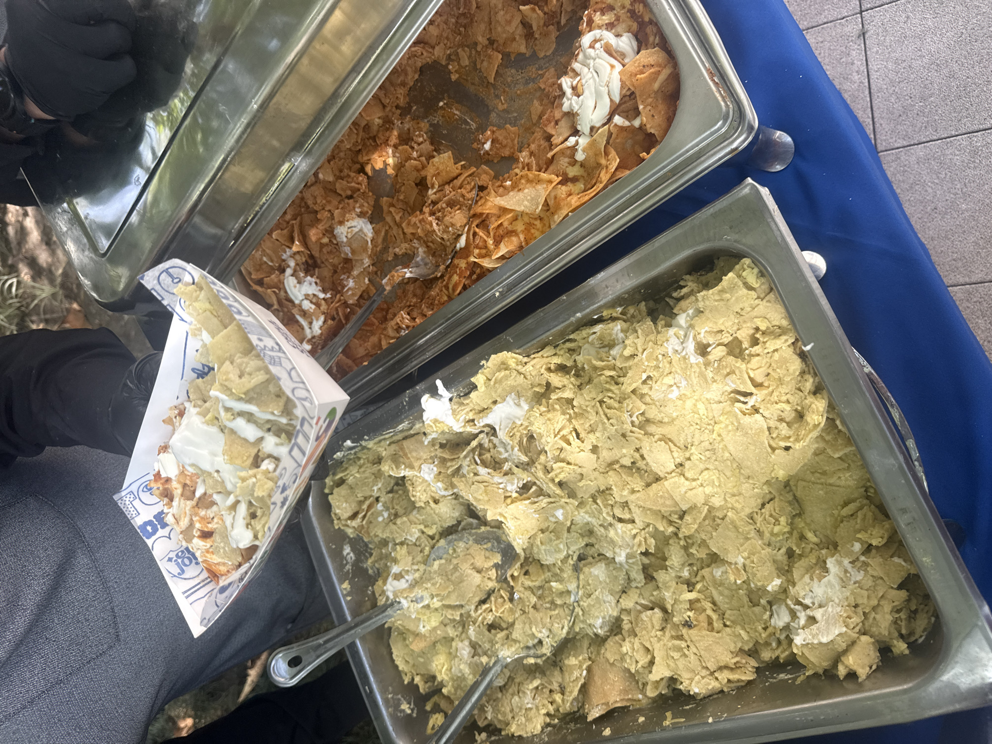 This media file captures a food service scene, likely outdoors or in an open-air event setting in Mexico City, Mexico. The central focus is on two large metal chafing dishes or steam table pans filled with chilaquiles, a traditional Mexican dish made from fried tortilla pieces often simmered in salsa.
The upper chafing dish contains chilaquiles prepared with a reddish sauce, indicative of a red salsa, with several dollops of white crema (Mexican sour cream) spread throughout. A serving utensil is visible within this pan. The lower chafing dish holds chilaquiles with a yellowish or light green hue, suggesting a green salsa preparation, also generously topped with white crema, and containing serving spoons.
In the foreground, a person, partially visible, holds a white paper food container filled with a mixture of both red and green chilaquiles, generously topped with white crema. The person's grey pants and a hand wearing a black glove are visible on the left. The paper container features blue graphics and text, with legible parts including "jör" and fragments like "DR" and "POP," likely indicating branding.
The chafing dishes rest on a vibrant blue tablecloth. The background shows a paved or tiled floor surface. The natural lighting suggests it is daytime. The overall scene depicts a casual food distribution point, offering fresh chilaquiles to attendees.