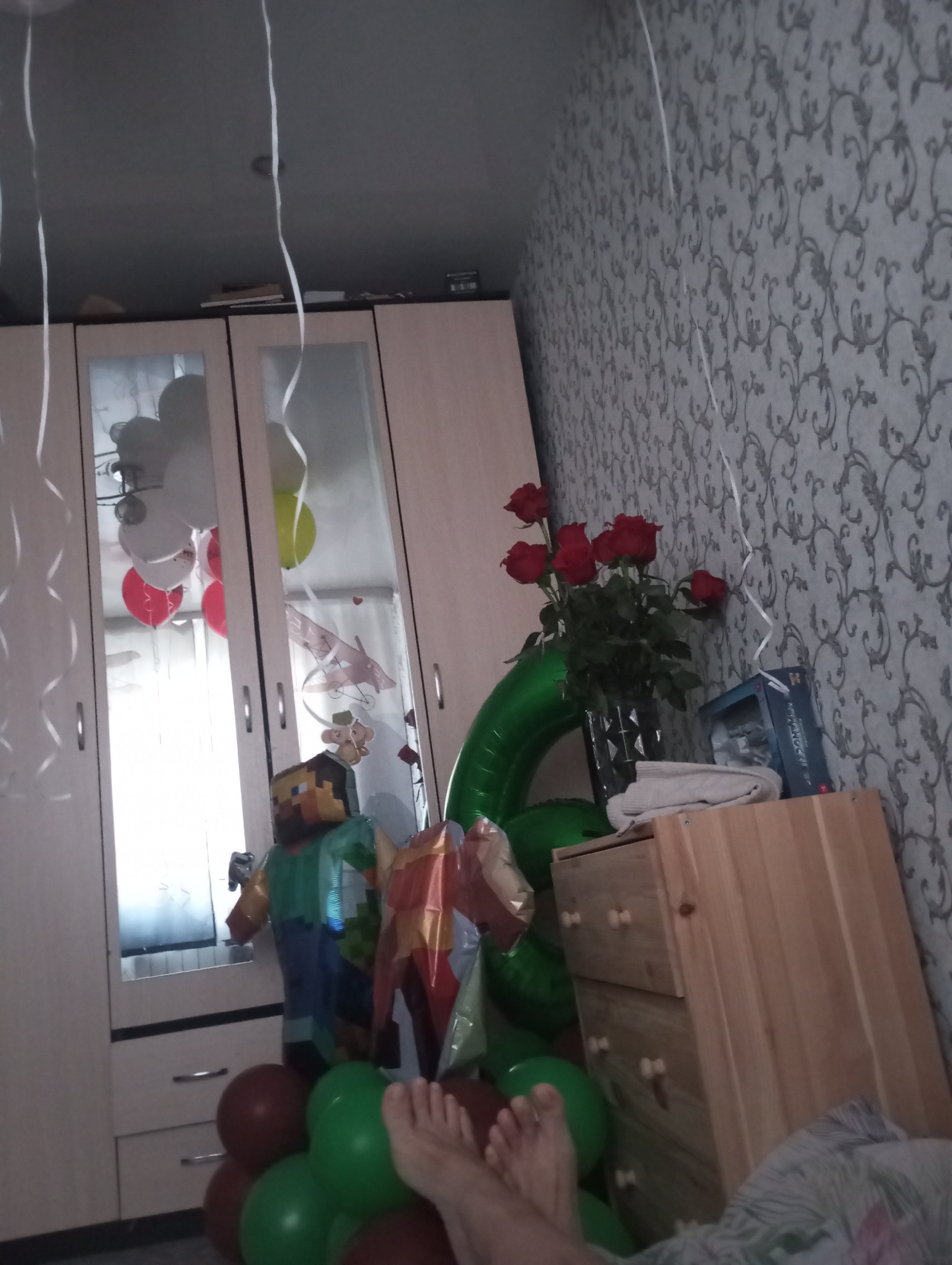 The photograph appears to be taken indoors, in a room decorated for a celebration. The wallpaper has a patterned, floral design. A light-colored wardrobe with mirrored doors is present, reflecting balloons of various colors (white, red, yellow) attached to strings. Additional balloons (red, green, maroon) are clustered near the floor. A balloon shaped like the number "6" is visible. A bouquet of red roses is in a vase, positioned on a wooden chest of drawers. Beside the roses is a box, possibly containing a toy or gift. A Minecraft-style figure is present, alongside a foil balloon. A person's bare feet are visible in the foreground. Given the question's constraints, the location is presumed to be in Kamensk-Uralsky, Russia.
