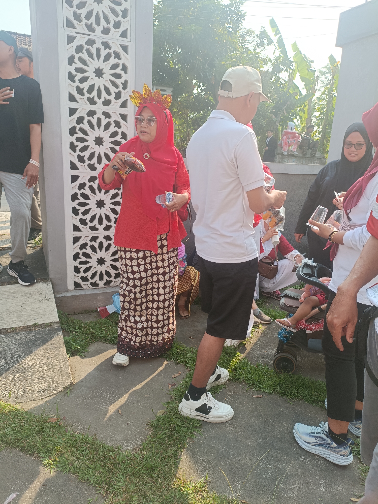 In the city of Sleman, Indonesia, on a sunny day, a group of people are gathered outdoors. In the foreground, a woman wearing a red hijab, a red patterned top, and a batik skirt stands holding a chocolate bar and a plastic bottle. To her right, a man in a white polo shirt and black shorts, wearing a beige baseball cap, holds a plastic bag with bottles. Further to the right, a woman in a black hijab and glasses is visible, and partially obscured, a person in a white shirt with red accents. A stroller with a child in it is positioned between the man and the woman in the black hijab. In the background, a decorative wall with geometric patterns is visible, along with trees and vegetation. The ground is paved with concrete and patches of grass. The overall scene depicts a casual gathering or event.