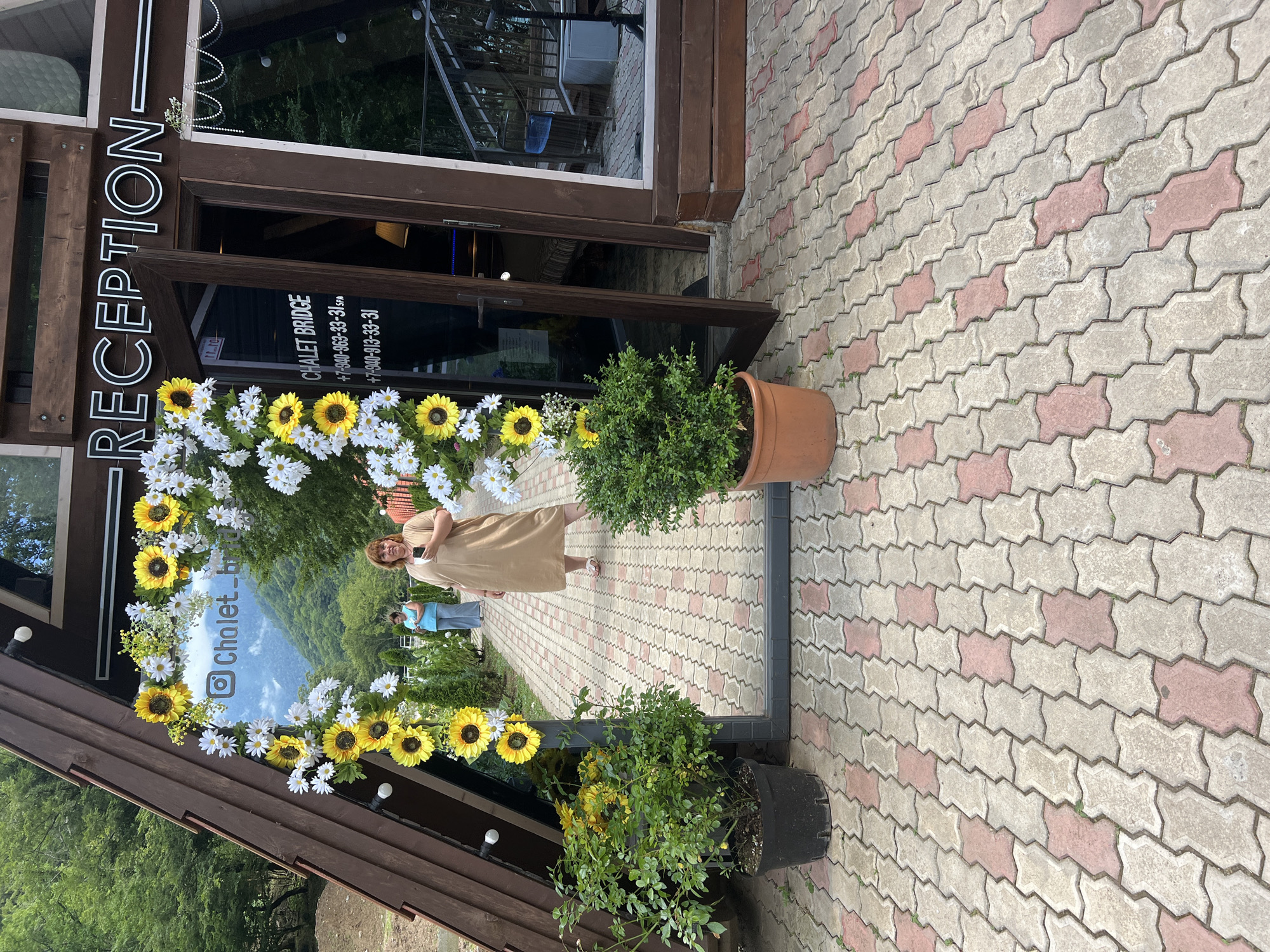 A woman stands in front of a building with a "RECEPTION" sign. She is wearing a light brown dress and holding a phone. In front of the building is a floral arch made of sunflowers and daisies. The ground is paved with interlocking bricks in red and gray. The reflection in the glass door shows a paved walkway, other people, and lush green trees and mountains in the background, suggesting an outdoor location with natural scenery. A potted green plant sits to the right of the floral arch. Another potted plant with yellow flowers is visible in the foreground. The time of day appears to be daytime, likely sunny given the lighting. The overall mood seems pleasant and welcoming. Visible text includes "RECEPTION" on the building and information on the glass door which includes "CHALET BRIDGE" and phone numbers. An Instagram logo and handle "@Chalet_br" are also visible on the floral arch.