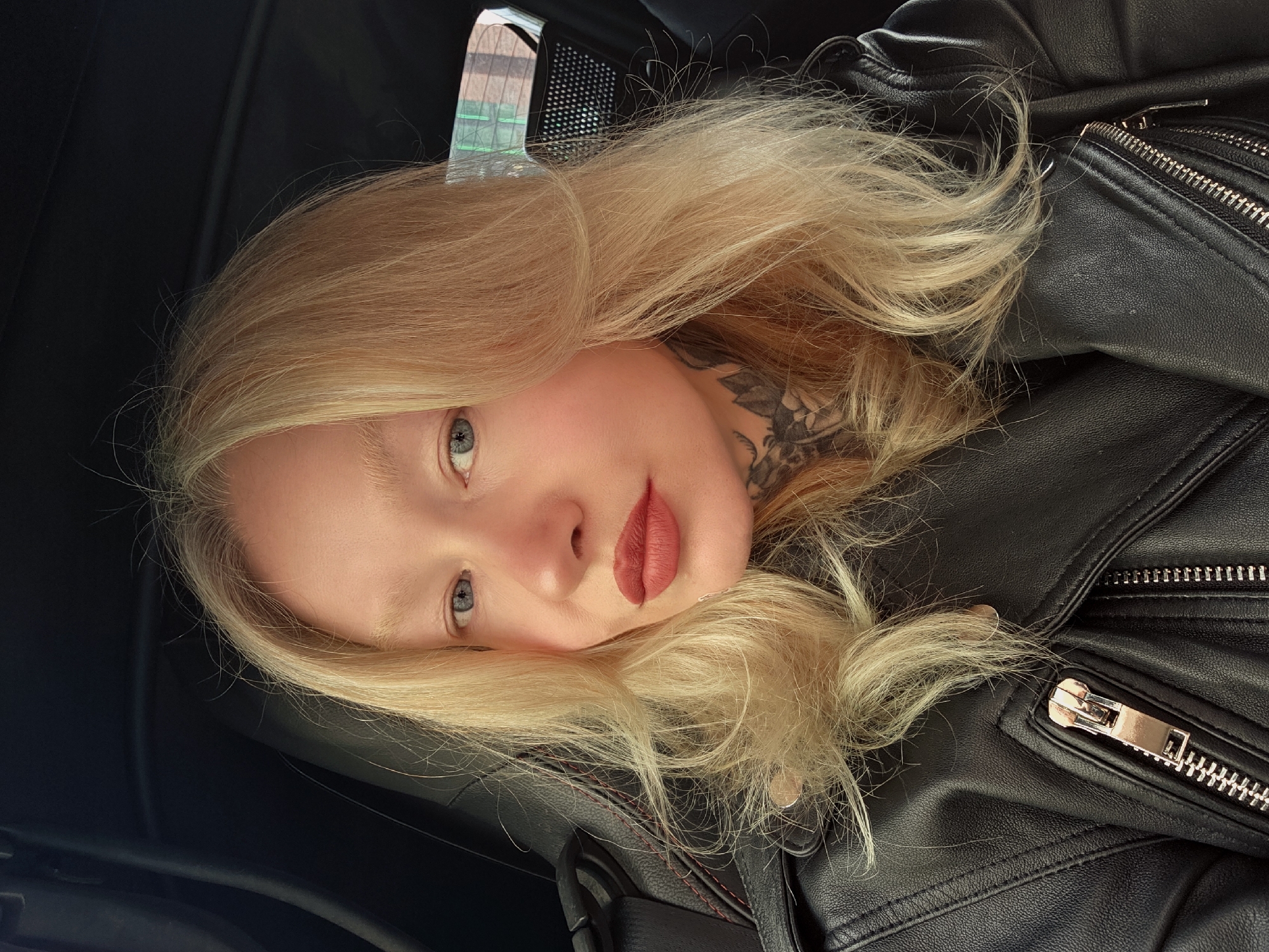 A fair-skinned individual with blonde hair is lying down, possibly inside a vehicle due to the dark interior. The person has a tattoo on their neck and is wearing a black leather jacket. The image may have been taken in Saint Petersburg, Russia, but there is no visual evidence in the photo to confirm this.
