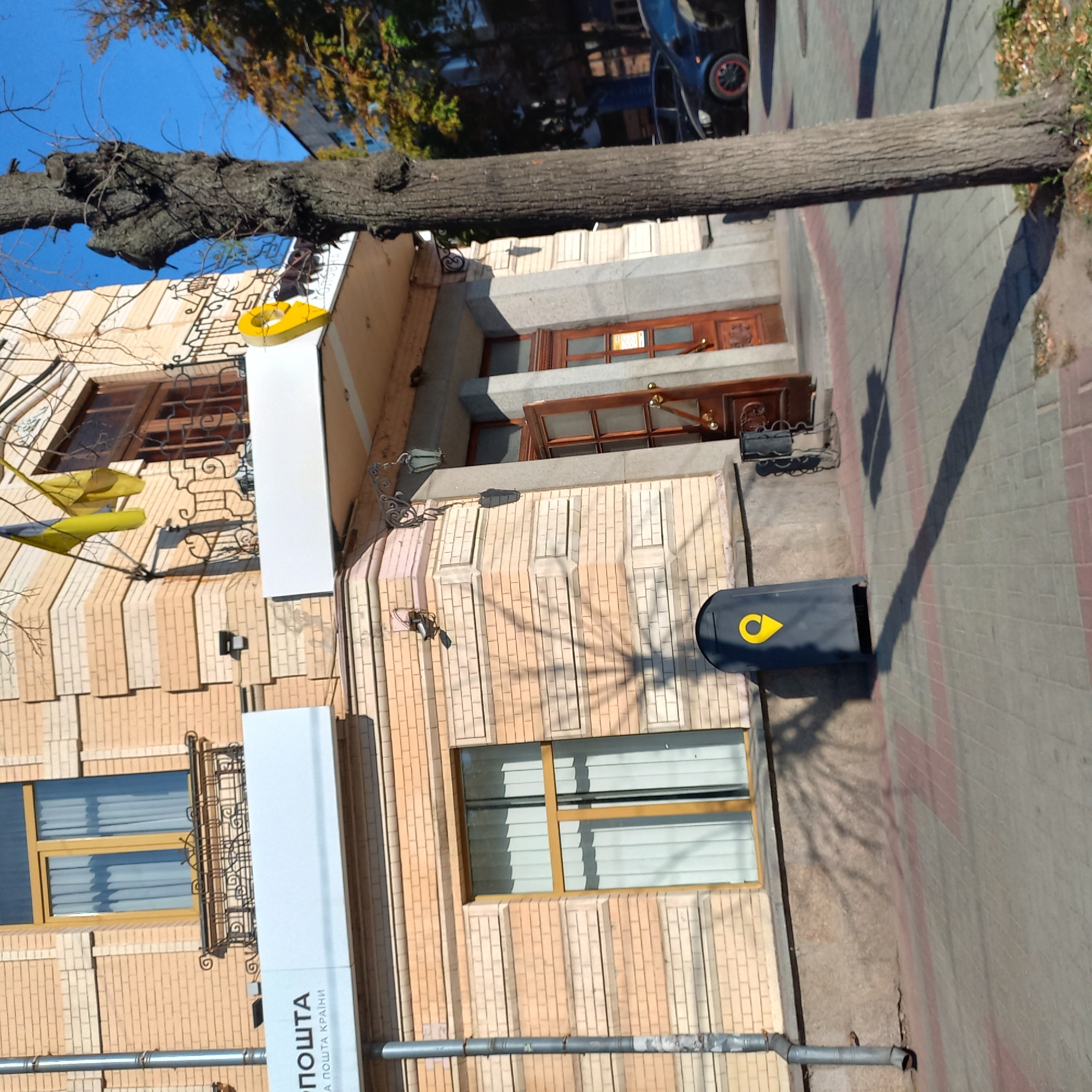 The image shows the exterior of a building with a brick facade in the city of Kropyvnytskyi, Ukraine. The building has several windows with yellow frames and white curtains. A metal drainpipe runs vertically along the left side of the building. In front of the building, there is a sidewalk paved with gray and pink tiles. To the left, there is a dark gray mailbox with a yellow location pin symbol. A large tree with rough bark stands in the foreground to the right, casting a shadow on the sidewalk. Ukrainian flags are visible on the upper facade of the building. There are also two cars parked on the street in the background. The scene appears to be a sunny day, as indicated by the strong shadows.