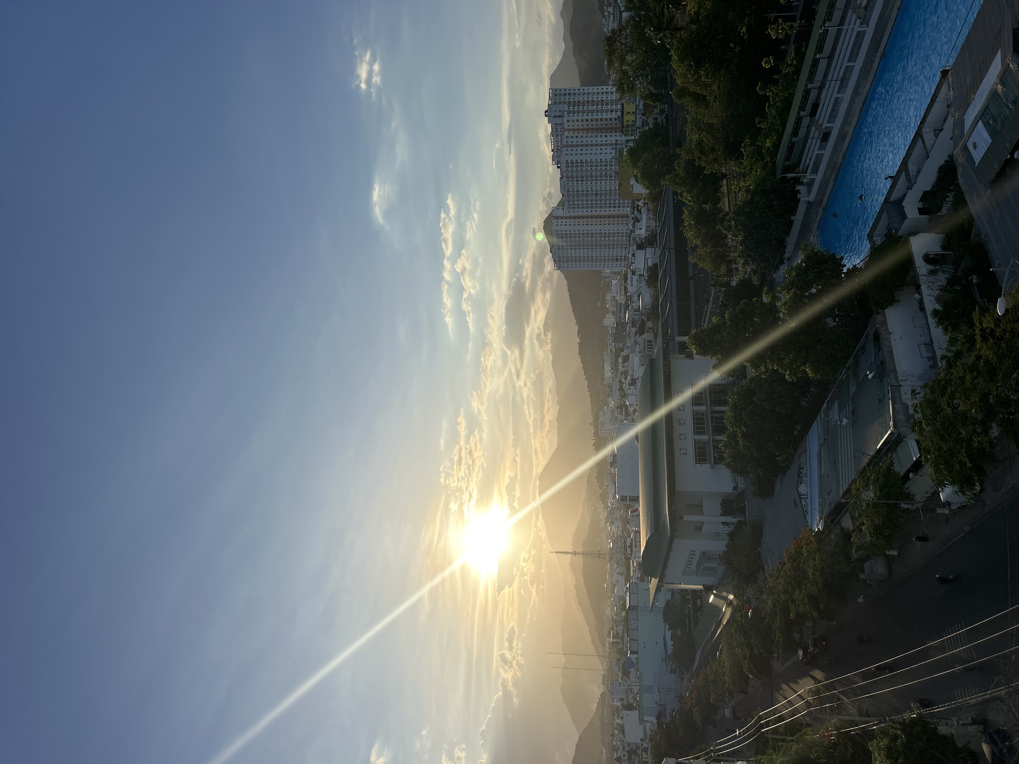 Here is a detailed description of the image:
The image presents an aerial view of Bac Nha Trang Ward, Vietnam, bathed in the warm glow of the setting sun. The sky occupies the upper left portion of the frame, displaying a gradient of blue hues interspersed with wisps of white clouds. The sun, a brilliant orb, is positioned near the horizon, casting long, luminous rays across the landscape and creating a pronounced lens flare.
Below the sky, a mountainous terrain is partially obscured by the haze of the setting sun, with tall buildings visible in the distance. The architecture blends residential structures with higher-rise apartment complexes.
In the lower right portion of the image, a series of buildings and structures emerge. A long, rectangular swimming pool filled with water is visible atop one of the structures. Roads, vehicles, and lush greenery contribute to the vibrant urban landscape. Power lines are visible.