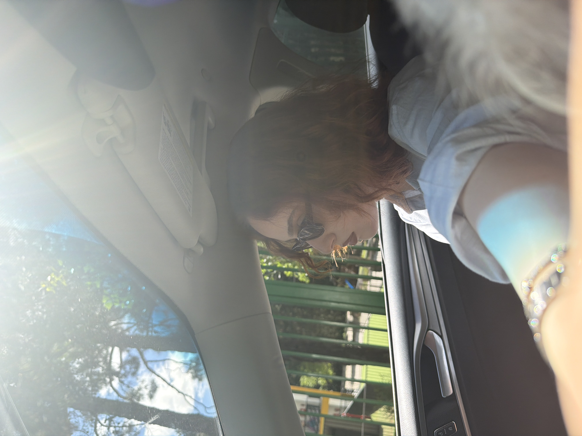 The image shows a person inside a car, likely in Tbilisi, Georgia, based on visual clues. The interior of the car is visible, including the sun visor, windshield, and door panel. The person, possibly a woman, has red hair and is wearing sunglasses. The exterior of the car, as seen through the window, shows greenery and a building, suggesting an urban or suburban area within Tbilisi. The overall impression is of someone sitting in a car, potentially parked or stopped, in a location within Tbilisi, Georgia.