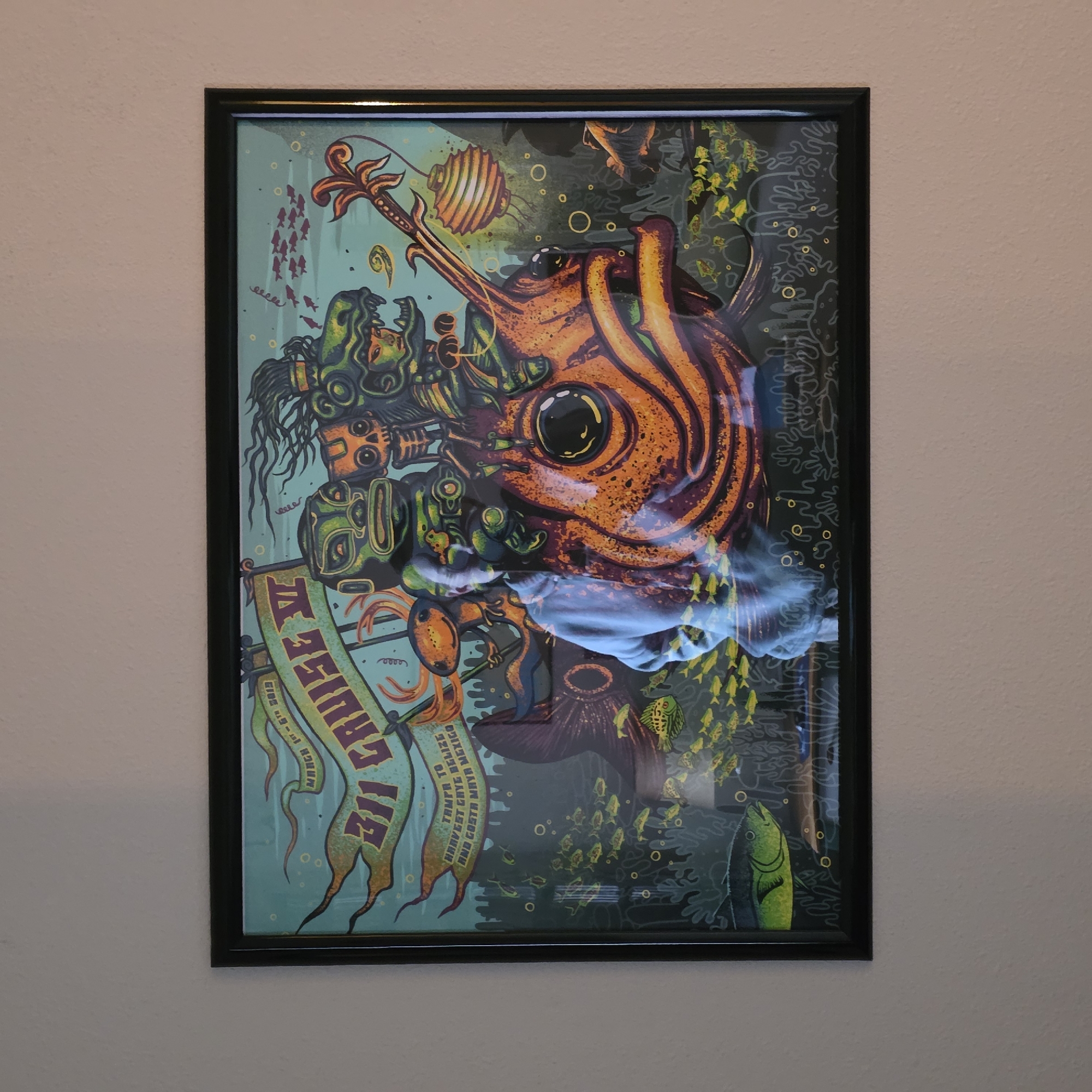 This media file shows a framed poster hanging on a textured, light-colored wall. The poster features a vibrant and detailed illustration with an underwater theme. The central subject is a large, orange, fish-like creature with a prominent eye and swirling patterns. Surrounding this creature are various stylized figures and elements, including what appears to be a mask, tentacles, and numerous small fish depicted in both red and yellow. There are also decorative elements like bubbles and what looks like coral.
A banner, predominantly green and yellow, is draped across the lower left portion of the poster. Visible text on this banner reads "311 CRUISE" and mentions "MARCH 1ST - 5TH 2013". Further text beneath this states "TAMPA TO HARVEST CAYE BELIZE AND COSTA MAYA MEXICO". The overall style is artistic and somewhat surreal, with a dark background contrasting with the brightly colored subjects. The lighting in the room suggests it is daytime, and the wall texture and frame indicate the poster is a physical object, likely a concert or event poster. The presence of reflections on the poster's surface suggests it is protected by glass within the frame. There are no visible people or overt emotional expressions conveyed in the artwork itself.