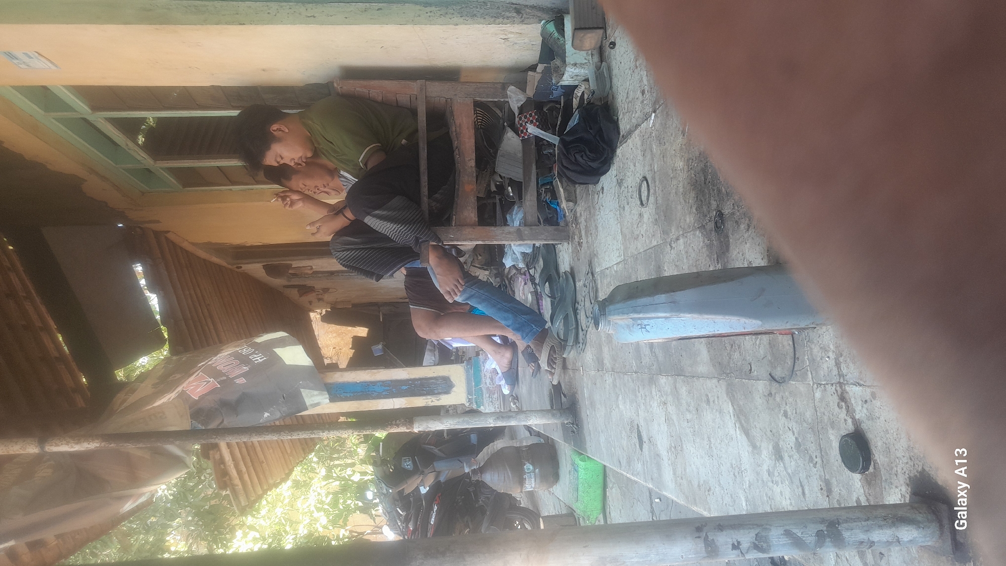 Two young men are sitting on a wooden chair under a dilapidated roof in Hadipolo, Indonesia. The man on the right, wearing a green t-shirt, is looking down at his phone. The man on the left, wearing a grey t-shirt and black and white checkered pants, is also looking at his phone and smoking a cigarette. In the background, a motorcycle is parked next to a large water jug. The ground is paved with concrete tiles, and various objects, including tools and discarded items, are scattered around. The building appears to be made of brick and stucco, with a light blue trim around the windows. Sunlight streams in from the left, casting shadows on the ground. The overall scene depicts a casual, informal setting, likely a workshop or a roadside stall.