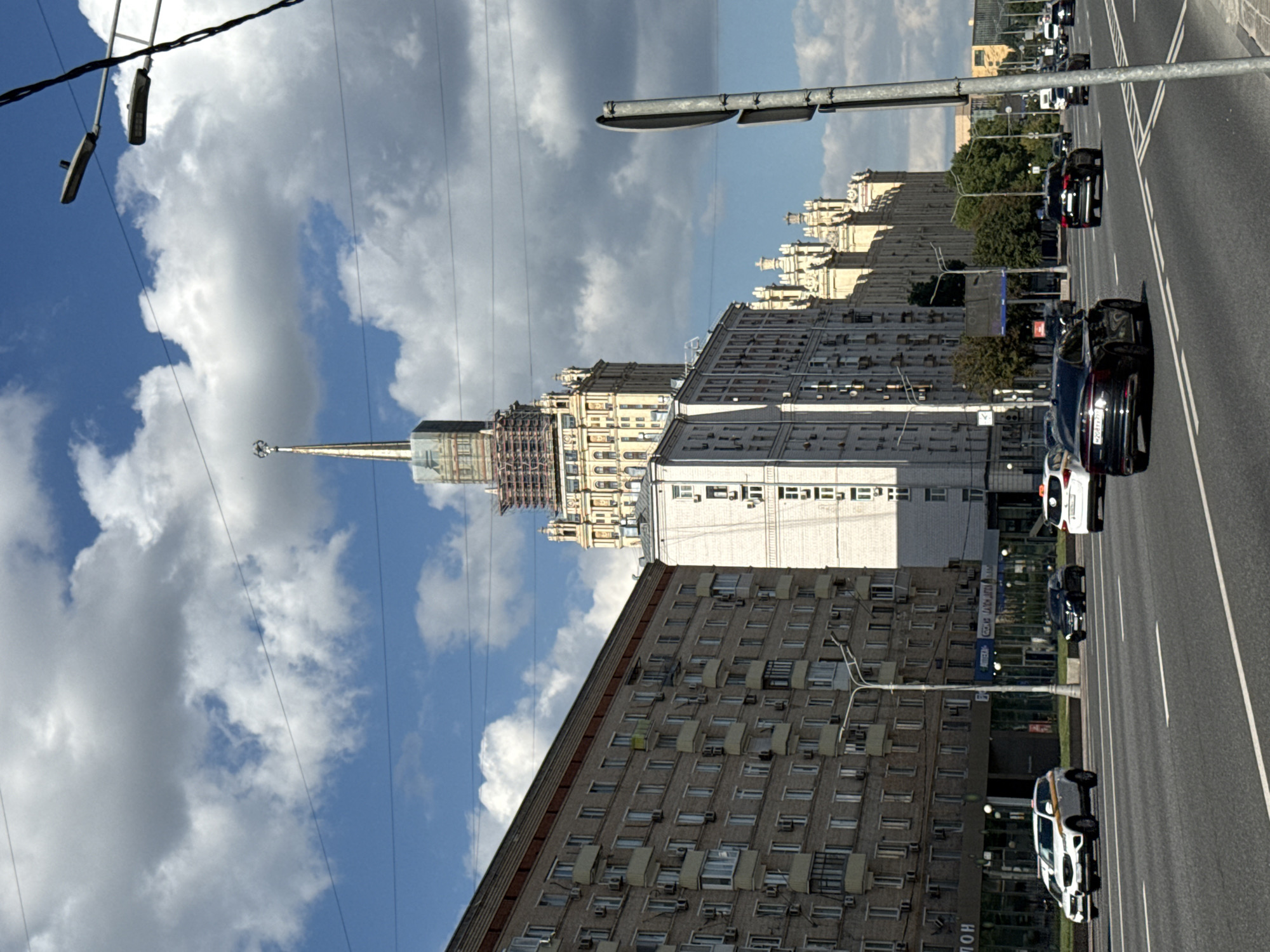 The image depicts a street scene in Moscow, Russia, with a prominent view of the Stalinist skyscraper, Kudrinskaya Square Building. The building's distinctive spire and ornate facade are visible, with scaffolding suggesting ongoing renovation or maintenance work. The foreground shows a multi-lane road with several cars, including a white SUV, a black sedan, and a white taxi, driving in the same direction. The buildings lining the street are a mix of Soviet-era apartment blocks and more ornate architecture, characteristic of Moscow. The sky is partly cloudy with bright sunlight, indicating daytime. The overall impression is a dynamic urban environment.