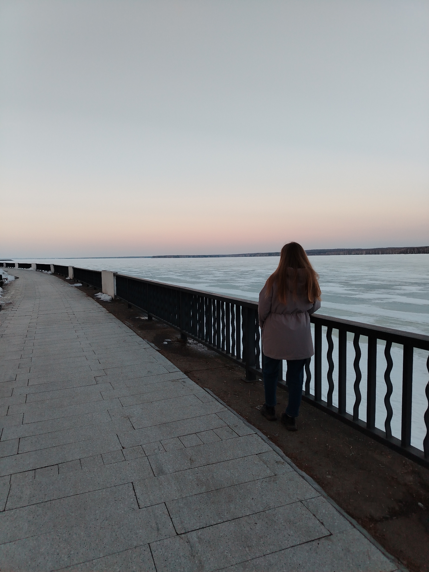 The image shows a woman standing on a stone-paved walkway, looking out over a body of water that appears to be partially frozen. She is wearing a gray jacket and blue jeans, and her back is to the viewer. A dark metal railing separates the walkway from the water's edge. In the background, a line of trees is visible on the far shore. The sky is light with a gradient of pale orange and blue hues, suggesting either sunrise or sunset. The scene is likely located in the city of Chaikovsky, Russia.