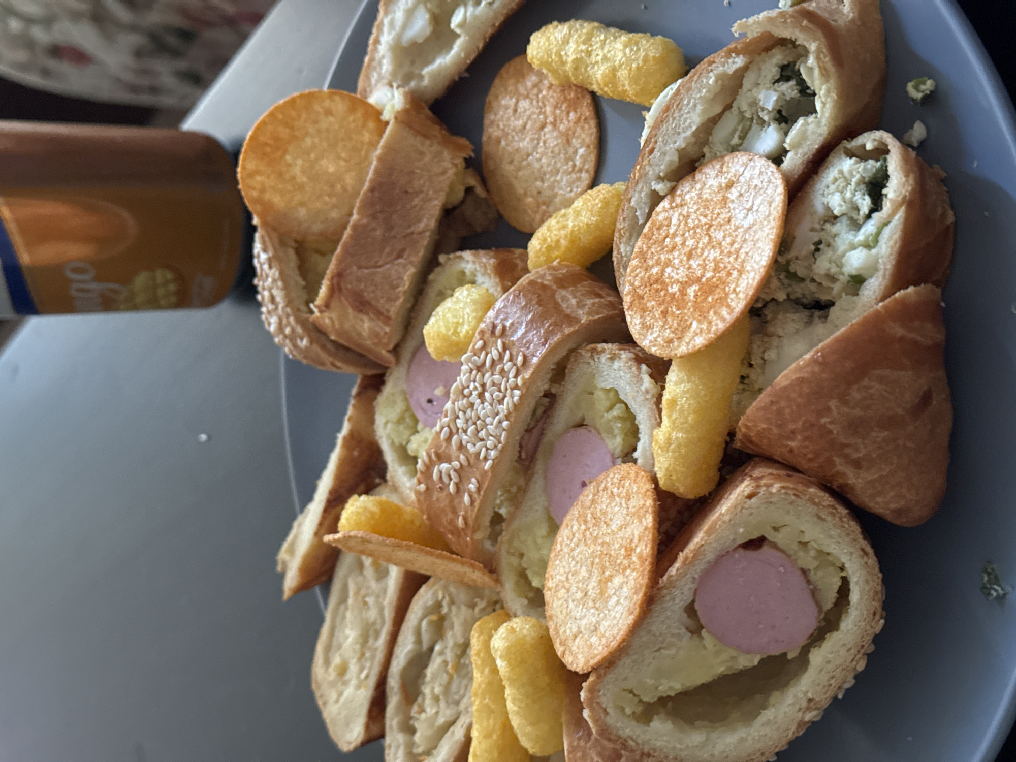 This media file shows a close-up, overhead view of a plate filled with food. The subjects are various food items, including pieces of bread with fillings, potato chips, and puffed corn snacks. Some of the bread pieces appear to be stuffed with what looks like mashed potatoes, a pink sausage, and a mixture of egg and green vegetables. The overall presentation is informal, suggesting a casual meal or snack. The lighting appears to be from an indoor source, casting soft shadows and highlighting the textures of the food. There are no visible people or a clear indication of the time of day or weather. The setting seems to be a domestic environment, possibly a kitchen or dining area, indicated by the presence of a gray plate and a portion of a bottle or can in the background. The bottle has some text that is partially visible, reading "mango" and "go" in a stylized font, suggesting a beverage. There are no other discernible location cues pointing to Krasnoyarsk, Russia. The food itself looks appealing and colorful, with the golden hues of the chips and bread contrasting with the pink of the sausage and the green elements in the filling.
