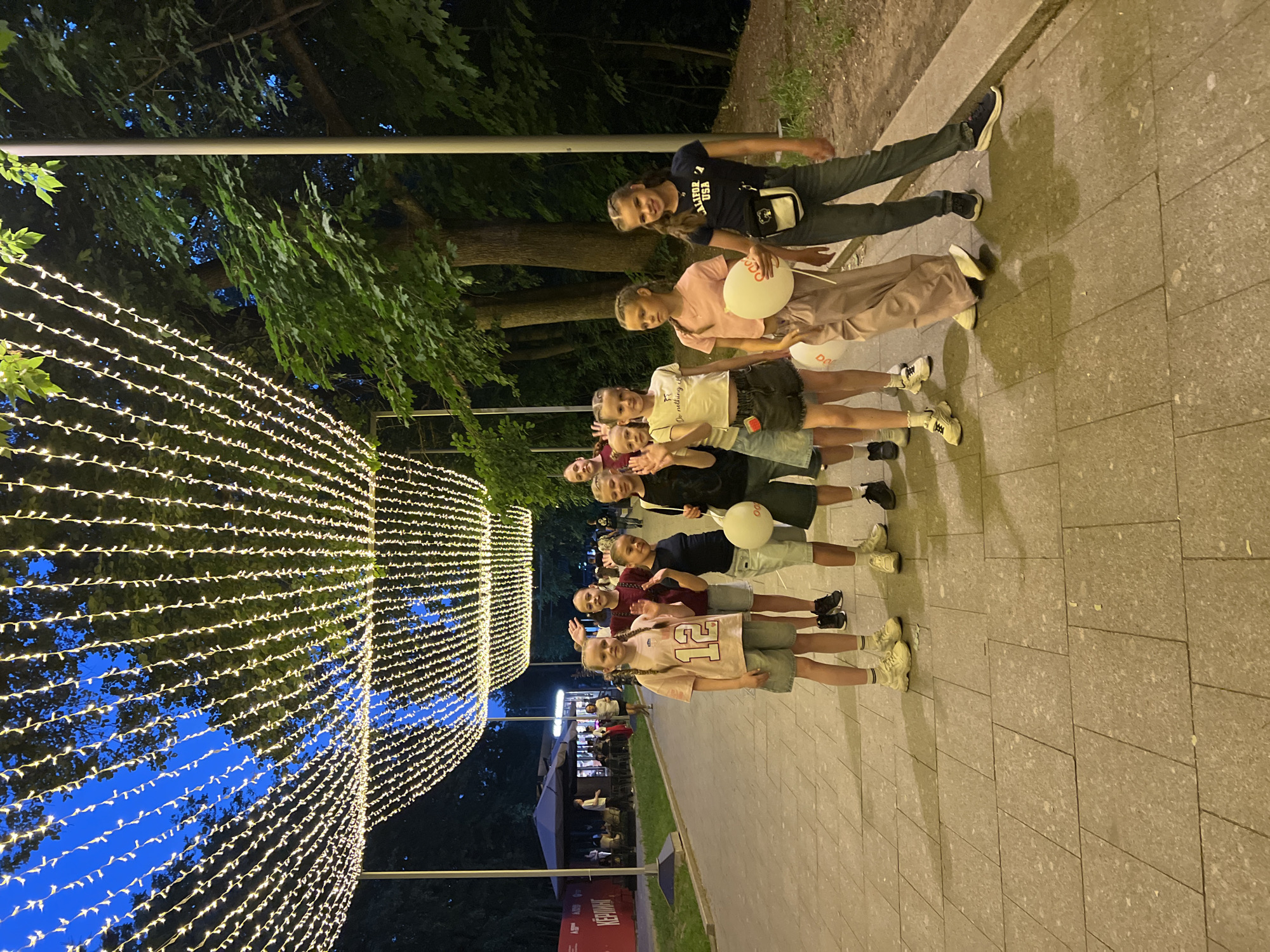 A group of nine children, appearing to be between 8 and 15 years old, are standing on a paved walkway in what is likely a park or public square in Moscow, Russia, under a canopy of decorative string lights. The sky above is a deep blue, indicating dusk or early evening. The children are dressed casually in t-shirts, shorts, and jeans. Several of them are waving, and two are holding white balloons. The children are positioned in a loose line, with the youngest-looking child in the front left, and older children towards the right. Behind the children, there are trees with green foliage, and further in the background, there are outdoor seating areas and possibly a cafe or restaurant. The overall atmosphere appears to be festive and relaxed, suggesting a social gathering or outing.