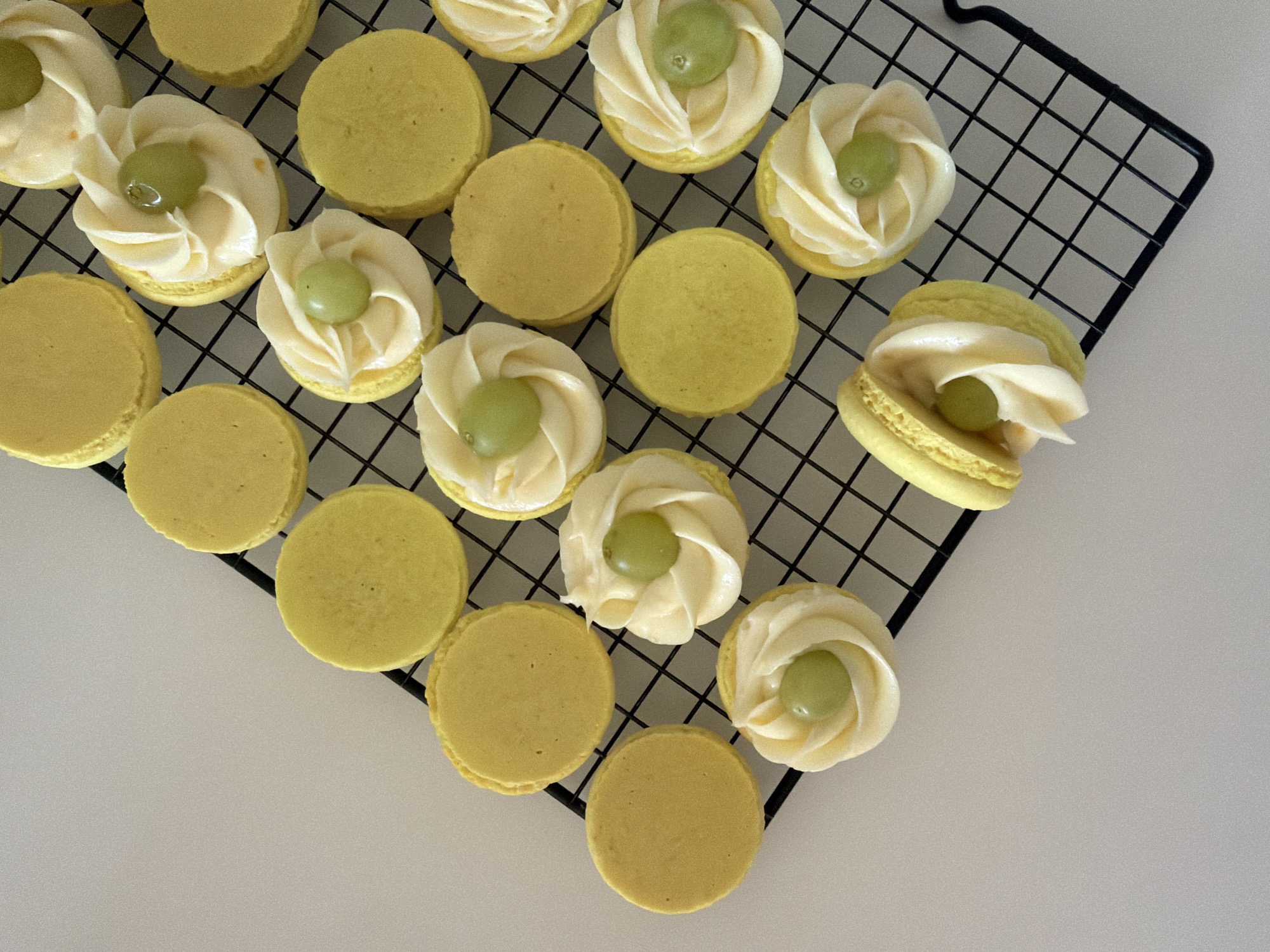 The image shows an array of yellow macarons on a black wire cooling rack. Some of the macarons are plain, while others are decorated with a white frosting swirl and a single green grape on top. The cooling rack rests on a white surface. The scene does not appear to be in Tel-Aviv, Israel, nor does it depict people or actions. The environment is a simple, indoor setting.