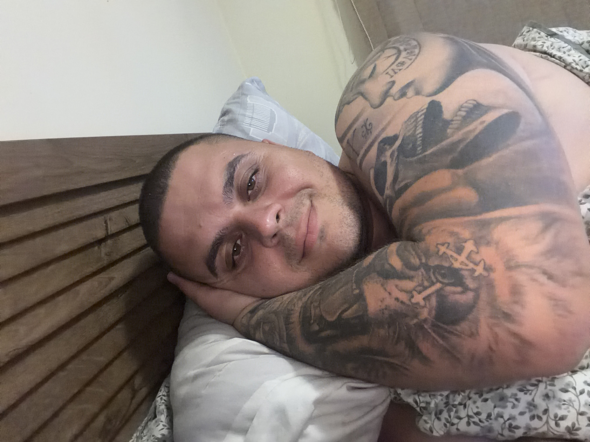 The image features a man lying in bed, looking directly at the camera with a slight, contented smile. His head is resting on a light-colored pillow, with his right arm bent and positioned beneath his head. The setting is clearly a bedroom, indicated by a dark wooden slatted headboard behind him and a patterned blanket with a floral or leaf design covering his left shoulder.
The man's right arm is extensively covered with tattoos, forming a full sleeve. On his upper arm, there is a detailed tattoo that appears to be a face or skull, possibly wearing a hat, along with what looks like partial Roman numerals or text, including characters that resemble "IASV" or "IASVI". Further down his forearm, a prominent tattoo of a large feline, possibly a lion or tiger, is visible. Superimposed over this animal tattoo are two distinct cross-like symbols, rendered in a lighter tone; one appears more ornate, while the other is simpler. Additional intricate designs and partial script, such as "XL," are also integrated into the sleeve.
The lighting in the room is soft and even, suggesting an indoor environment, potentially during the morning or evening. The man's expression conveys a sense of calm and relaxation. The context provided indicates this scene is in Shumen, Bulgaria.