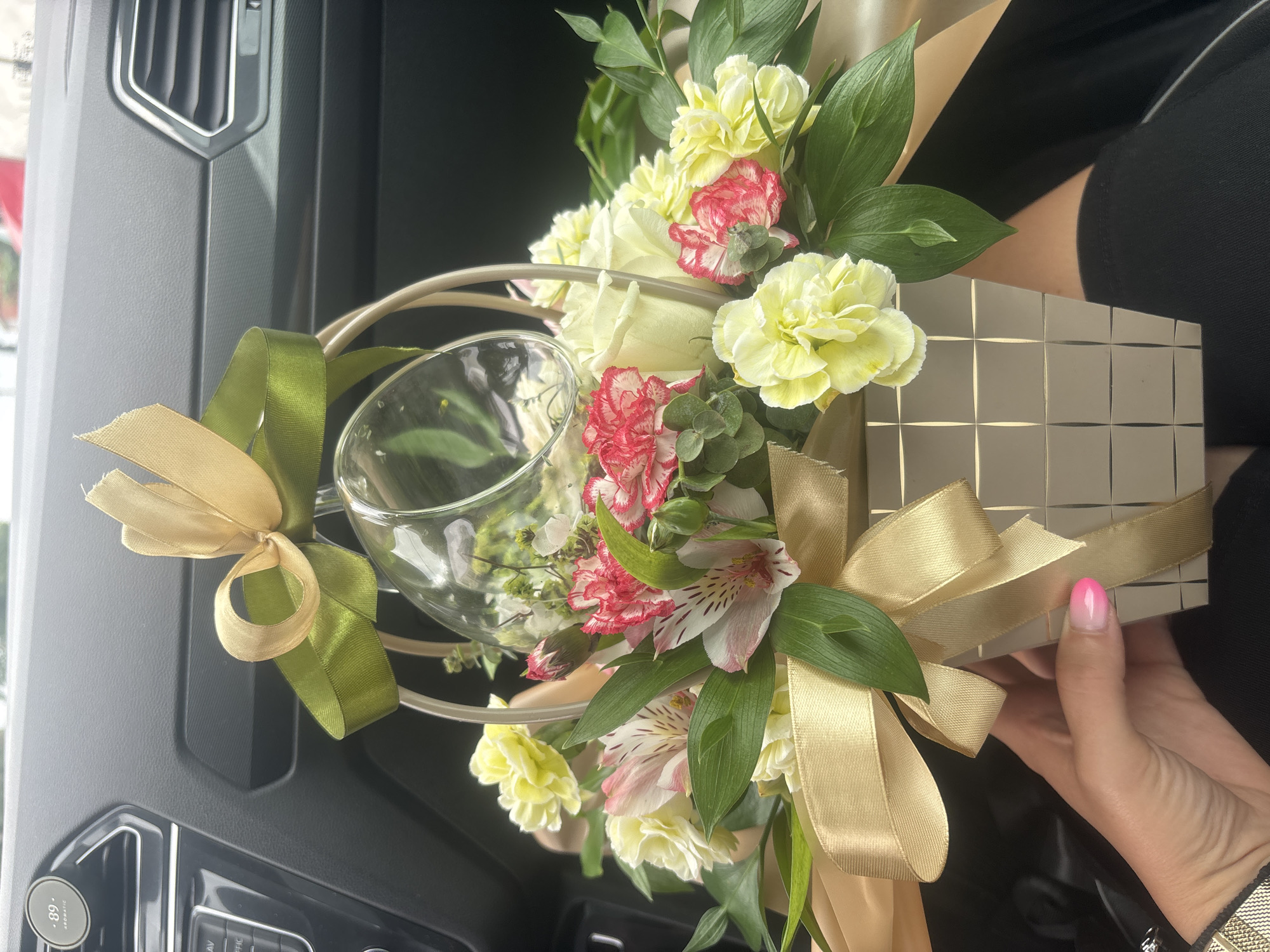 A hand with pink and white gradient fingernails holds a square, light-colored gift box filled with a floral arrangement, partially obscuring the box. The box is adorned with a gold ribbon tied into a bow. The floral arrangement includes white roses, yellow carnations, pink and white alstroemeria, and red-edged carnations, with green foliage. A clear glass vessel is inserted into the top of the arrangement. The arrangement is positioned inside a car, with the dashboard and air vents visible in the background, suggesting the car is parked in a location within the city of Burgas, Bulgaria. The lighting suggests an outdoor, daytime setting.