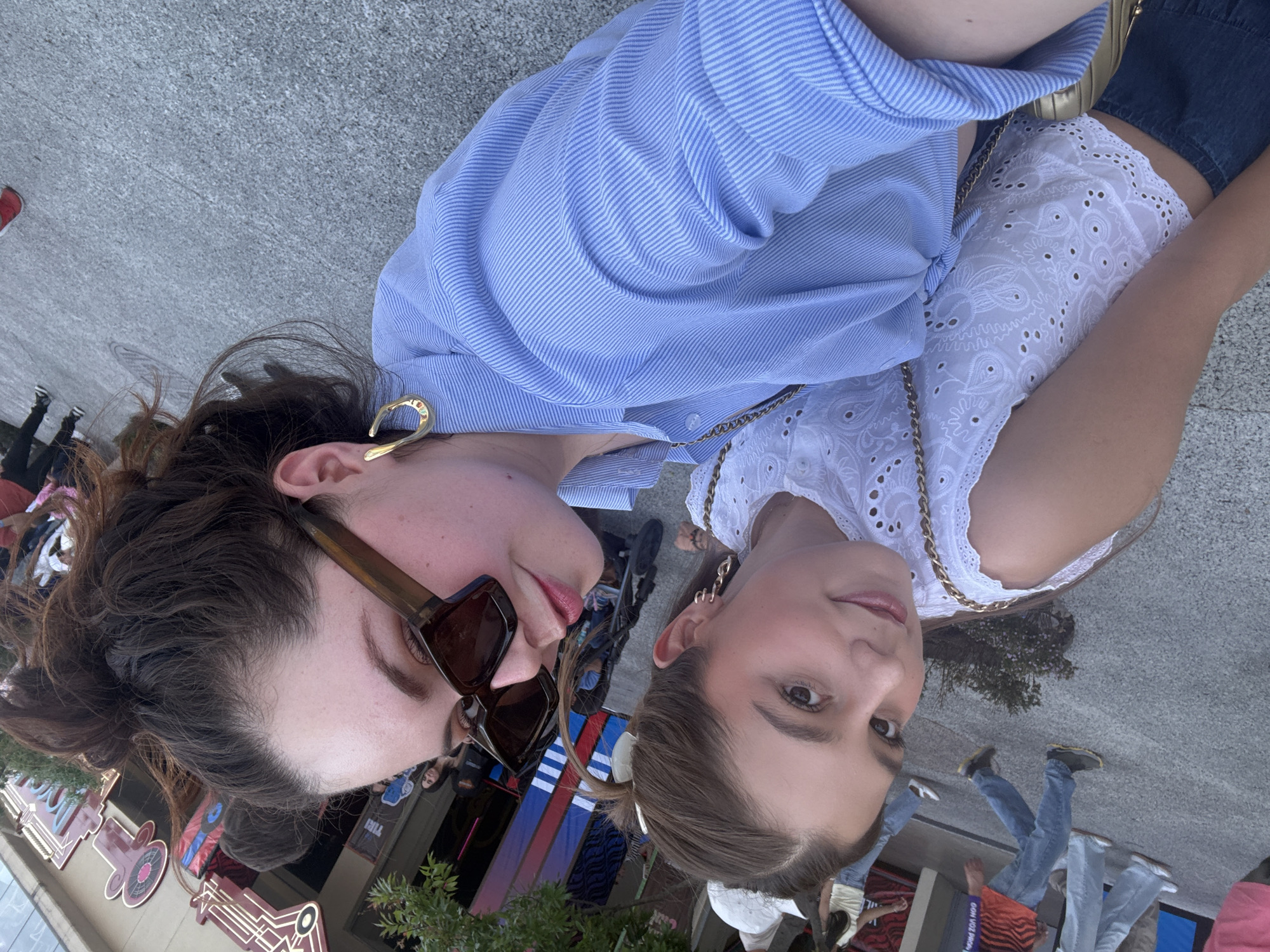 Two young women pose close together for a photo outdoors. The woman on the left has shoulder-length brown hair, large brown sunglasses, and a large gold earring. She is wearing a blue-striped collared shirt. The woman on the right has brown hair pulled back into a ponytail. She is wearing a white lace top with a gold chain necklace.
The background shows a street scene with pedestrians and a building façade with decorative elements. Political signs are visible in the background. The location is Mexico City, Mexico.