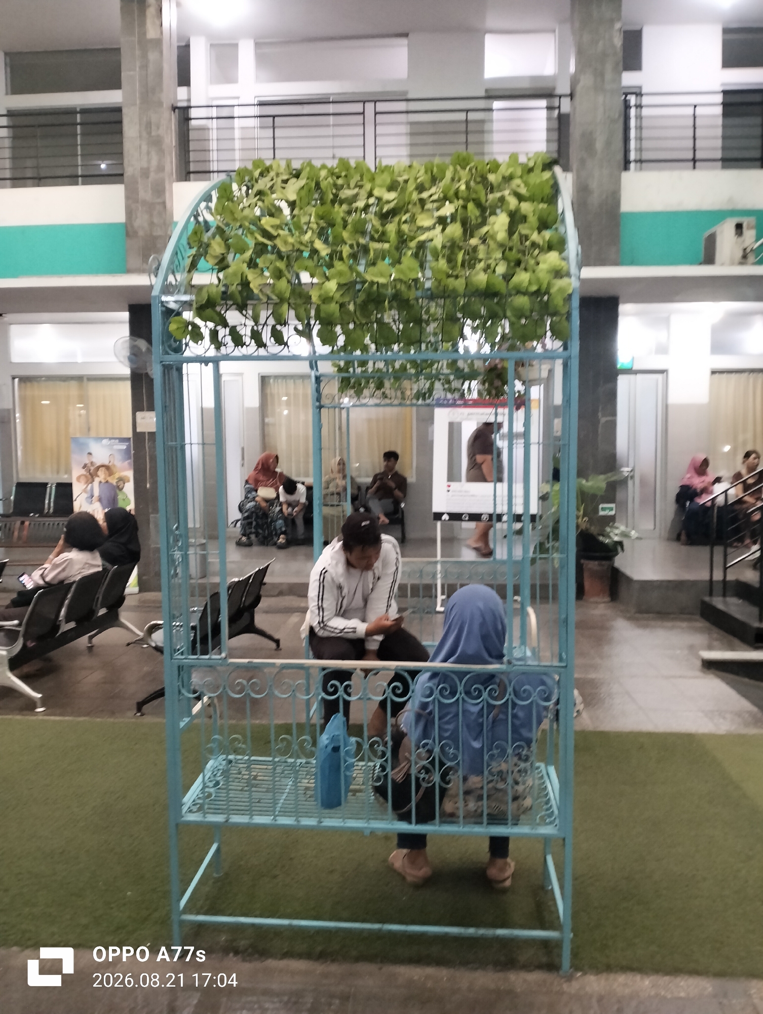 The image depicts a waiting area, possibly at a government building or a hospital in Semarang, Indonesia. In the foreground, a light blue ornate metal cage-like structure, overgrown with green plants, stands on artificial turf. Inside the cage, two individuals are seated. One person, wearing a blue headscarf, is facing away from the camera and is seated on the floor. Another person, wearing a white jacket with black stripes and dark pants, is seated to the left and is also facing away from the camera.
Beyond the cage, several other people are visible, seated on chairs in what appears to be a waiting hall. Some are looking at their phones, while others are conversing. In the background, there are windows, doors, and architectural elements consistent with a public building. The lighting suggests it might be indoors or late in the day. A poster featuring several people is visible on the left side of the image. The overall impression is one of a public space where people are waiting.