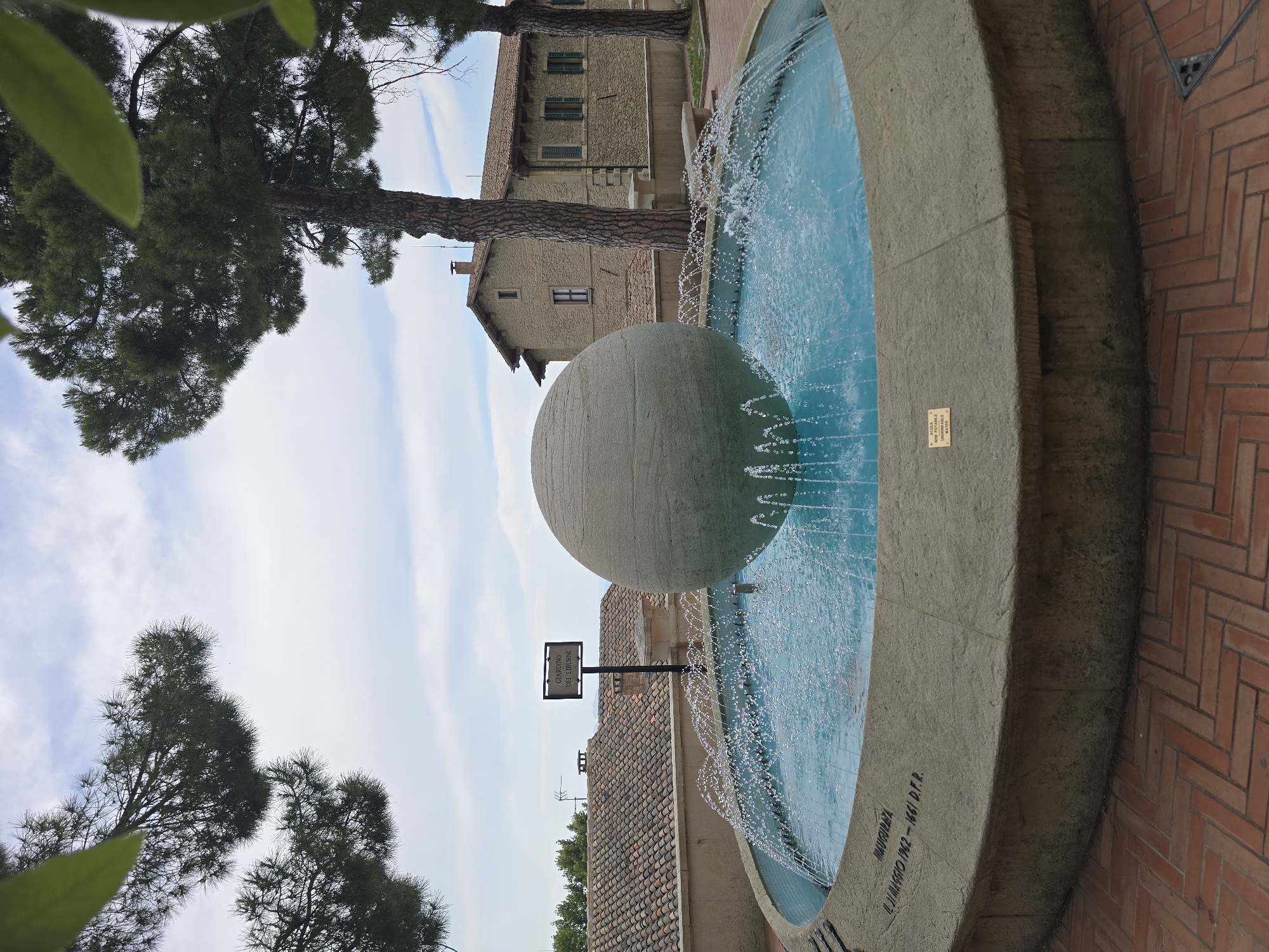 In the city of San Marino, San Marino, a large, spherical fountain centerpiece is prominently displayed within a circular fountain basin. The sphere, made of a textured, light-colored material, sits at the center of the basin, which is filled with bright blue water. Water streams upward from multiple spouts around the sphere, creating a dynamic visual effect. The fountain is constructed from a pale, concrete-like material, with visible inscriptions along its rim. One inscription reads "INAUGURATA IL 21 MAGGIO 1962 - 1661 D.F.R.", indicating its inauguration date. Another small plaque, mounted on the fountain's edge, states "ACQUA NON POTABILE - NON-POTABLE WATER" in both Italian and English. The fountain is situated on a patterned brick walkway. In the background, traditional buildings with terracotta tile roofs are visible, suggesting a historic urban setting. Evergreen trees with dark green foliage are present in the foreground and frame the scene. The sky above is partly cloudy, with white clouds scattered across a pale blue expanse.