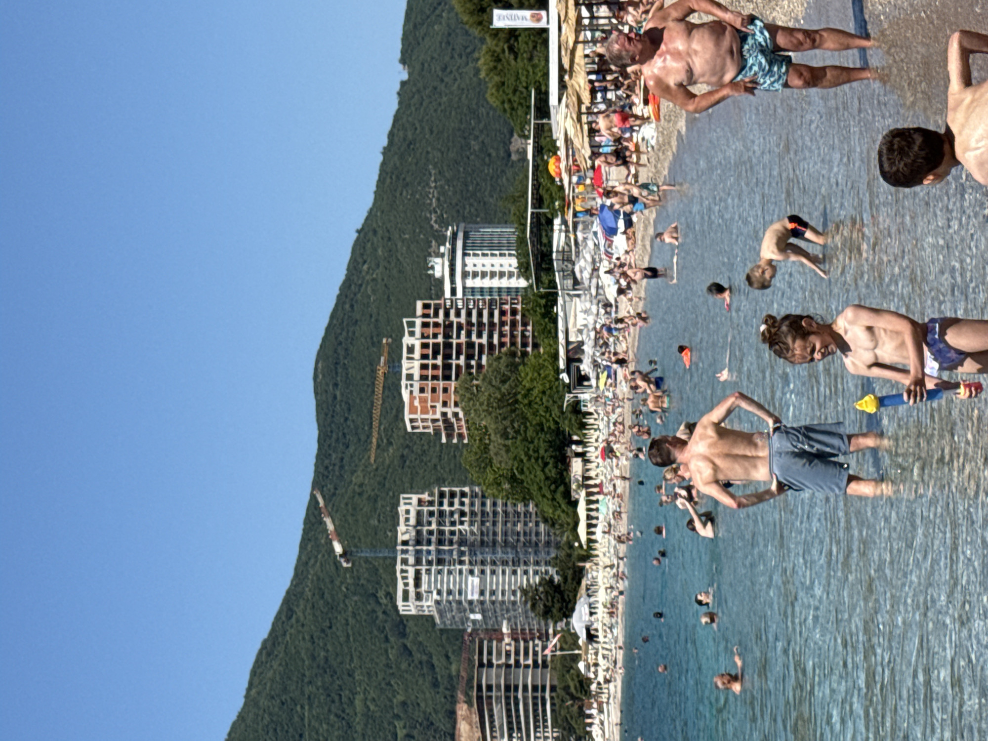 This image depicts a sunny, crowded beach scene in Boreti, Montenegro, with clear blue skies overhead. In the background, verdant mountains rise, partially obscured by several multi-story buildings, some appearing to be large concrete structures under construction with visible scaffolding, and others seemingly completed. Two prominent construction cranes are visible above the buildings and mountains; one is white and red, the other yellow.
The beach itself is densely populated with numerous individuals lounging on sunbeds under a mix of straw and fabric umbrellas. Many more people are in the clear, shallow, light-blue seawater, wading, swimming, or standing.
In the immediate foreground, from left to right:
* A young girl with dark hair tied in braids, wearing a patterned two-piece swimsuit, stands in the shallow water, holding a yellow and blue water toy, and looking slightly to her right with a faint smile.
* A young boy, shirtless and in dark swim briefs, bends over in the water, looking downwards.
* A man, viewed from behind, with a tanned back and wearing blue swim shorts, stands in the water with his hands on his hips.
* To the far right, another man with a stocky build, wearing patterned blue swim shorts, stands in the water, facing left and looking towards the water.
Further back in the water, numerous other people are partially submerged, engaging in various water activities. An orange flotation device is visible in the water on the far left. The overall scene suggests a vibrant holiday destination undergoing concurrent development.