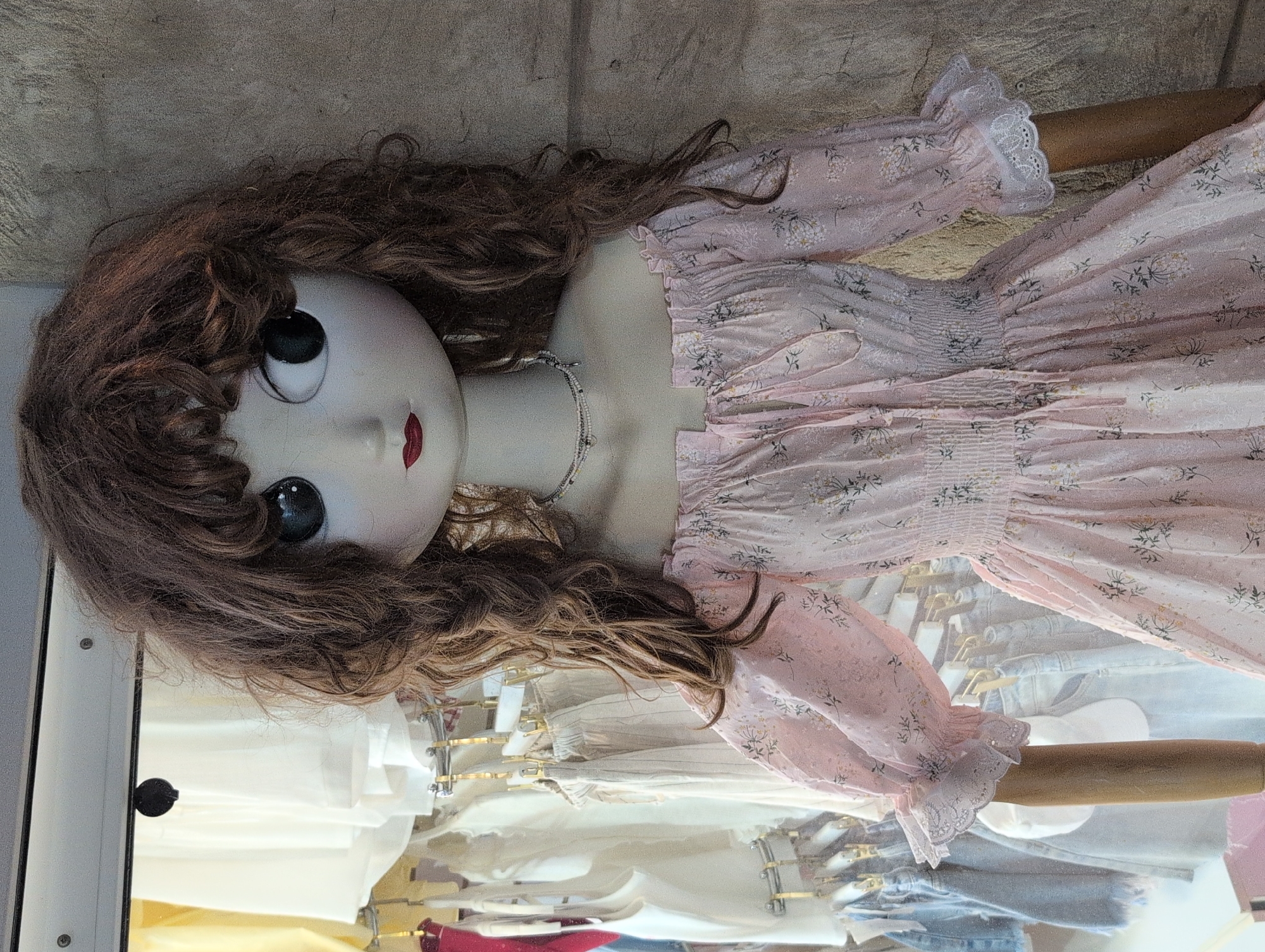 A mannequin with a large head and large eyes, wearing a pink floral off-the-shoulder dress, is displayed in what appears to be a clothing store. The background shows racks of clothing, including white tops and denim shorts. The mannequin's hair is brown and curly. The dress has elastic ruching at the waist and puffy sleeves with lace trim. The overall impression is of a display in a retail environment, possibly in a city like Gelendzhik, Russia, given the context of the image source. The lighting is bright, suggesting an indoor setting.