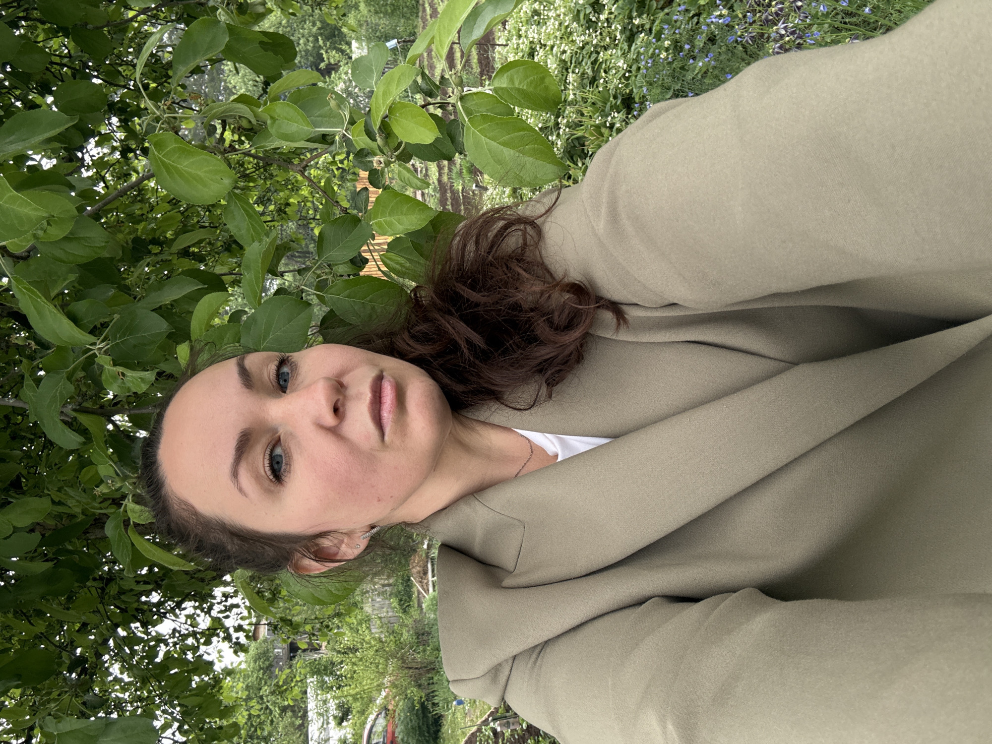 The image is a selfie of a woman with blue eyes and brown hair, wearing a light olive-green blazer over a white top. She appears to be outdoors, surrounded by lush greenery, including leafy branches in the foreground and a mix of green plants and possibly some wildflowers in the background. The background suggests a garden or park-like setting. The exact location within Chaikovsky, Russia cannot be determined from the image.