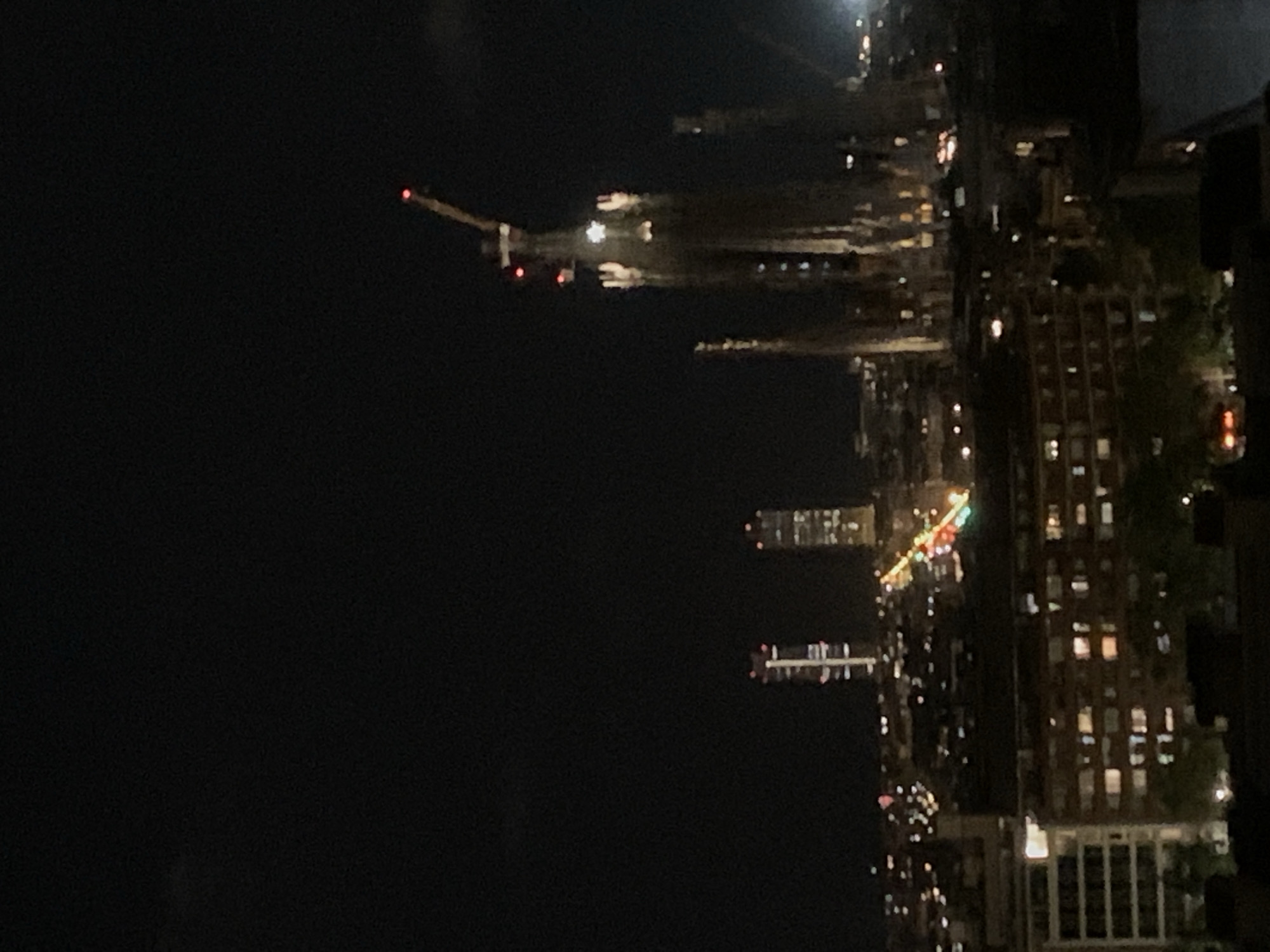 A low-angle, medium shot, taken at night, reveals a cityscape with identifiable architectural features.
To the right, rows of lit windows suggest a dense urban area. A large building is visible, illuminated with many windows. Streets are lined with lights, creating a visual texture.
To the left, a construction crane with a red light is visible against the night sky. This suggests ongoing development or construction activity.
In the central area of the frame, several skyscrapers and other high-rise buildings are visible. Their lights and illuminated outlines contribute to the overall nightscape.