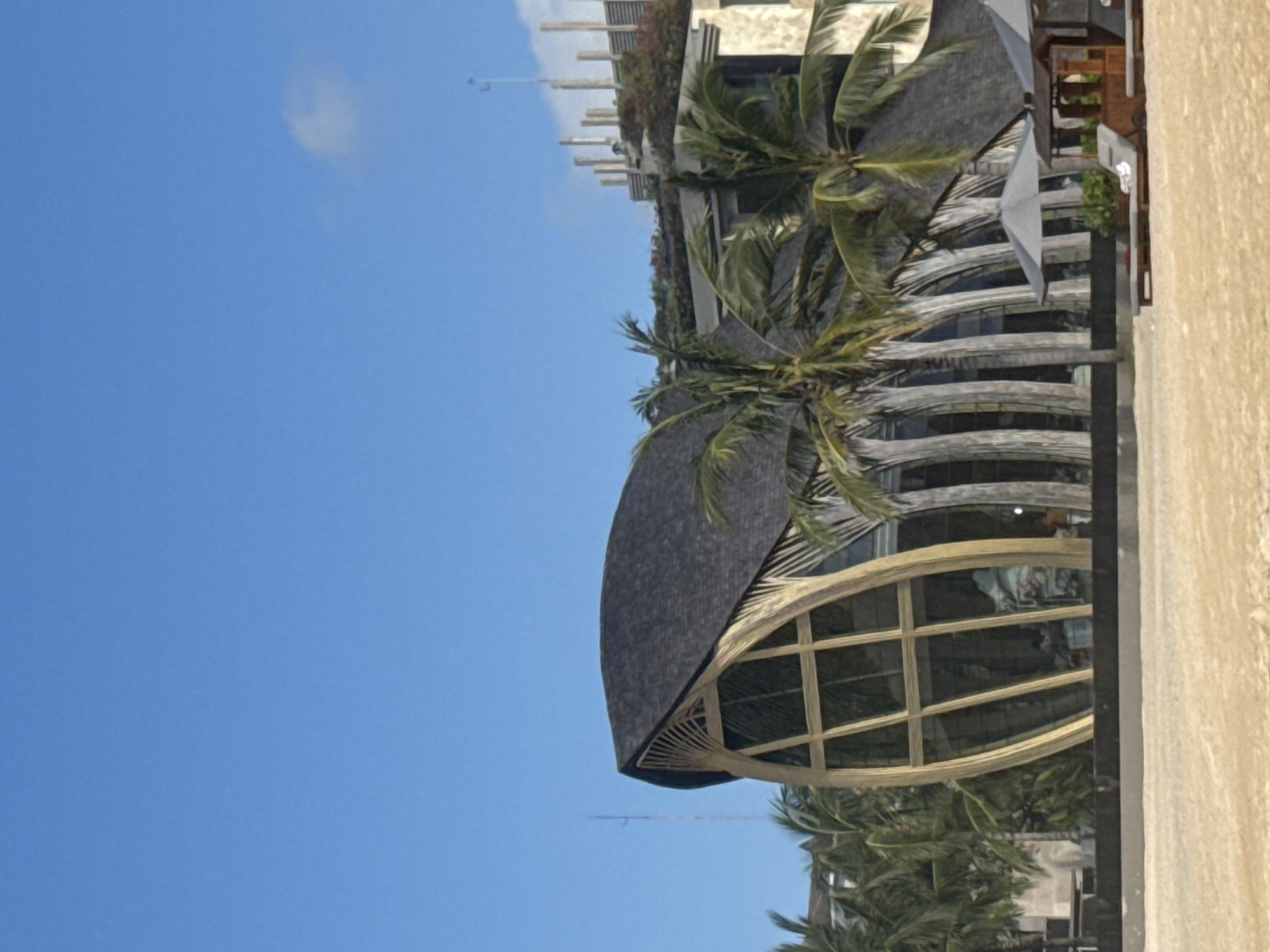 The image shows a building in Kuta Selatan, Indonesia. It has a curved, dark roof and a facade with large, arched windows. Palm trees are visible around the building. The sky is blue with a few clouds. Part of a light-colored wall is on the right side of the image.