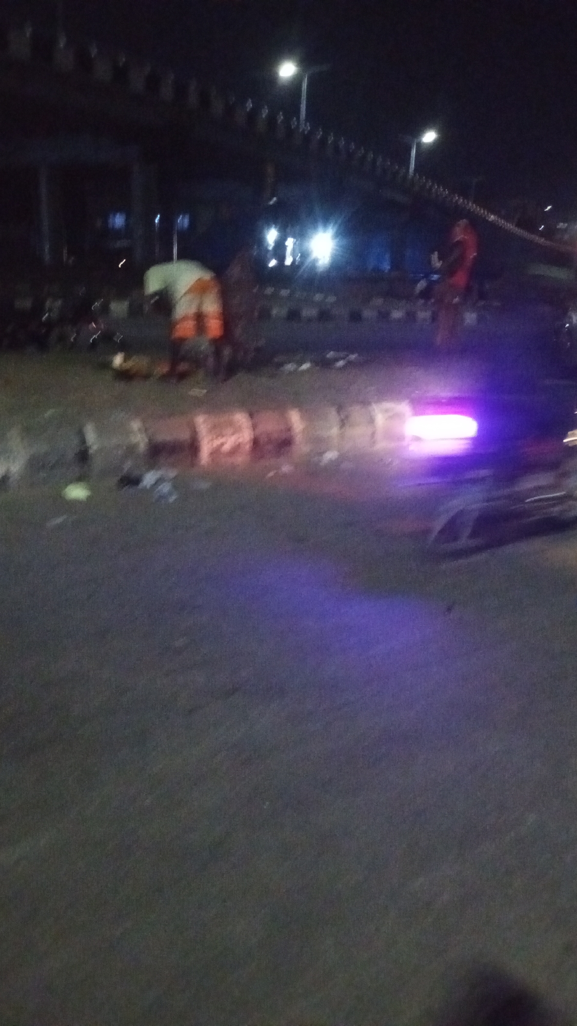 This media file captures a nocturnal scene on a street in Lokoja, Nigeria, likely depicting the aftermath of an incident. The time is clearly night, with the area illuminated by artificial light sources including streetlights mounted on overhead poles, bright headlights from a distant vehicle, and a distinct, intense purple/pink light source in the mid-ground.
In the left-midground, a person is visible, bent over, wearing a light-colored top (possibly white or light green) and orange shorts or trousers, seemingly attending to something on the ground. Further back and to the right, another individual stands, dressed in what appears to be a red or orange top.
The scene takes place on an asphalt road surface, which appears wet in sections, reflecting the various lights. A concrete barrier or divider runs across the mid-ground, and there is significant debris scattered on the road, particularly around this barrier and near the bending figure. The prominent purple/pink light source, possibly an emergency vehicle's light or a spotlight, is located on the right side of the road, behind the concrete barrier. Above, a dark, elevated structure resembling an overpass or bridge is visible, with its underside and railing illuminated by streetlights. No discernible text is visible within the image. The overall impression is one of a recent event or ongoing activity requiring attention, possibly an accident cleanup.